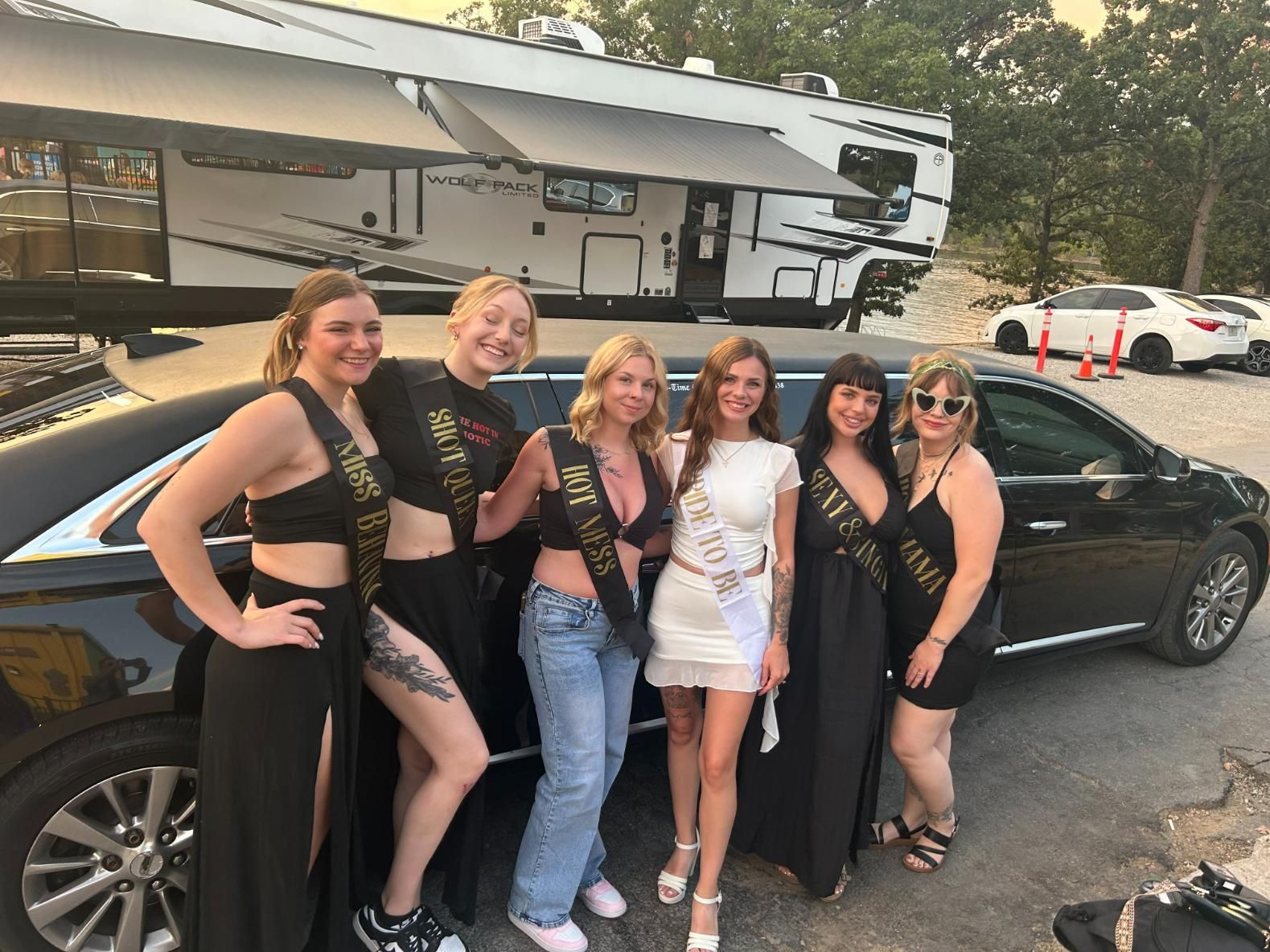 Six women in formal wear pose by a black limo and RV. They wear sashes, smiling in an outdoor setting.