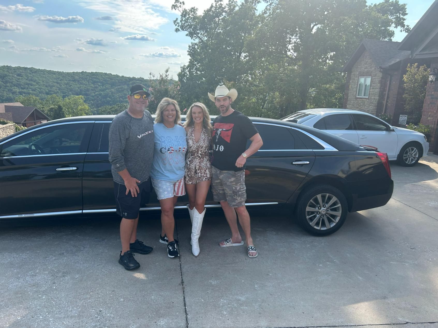 Four people pose by a black car; woman in boots, cowboy hat, patterned dress; other woman in stars/stripes.