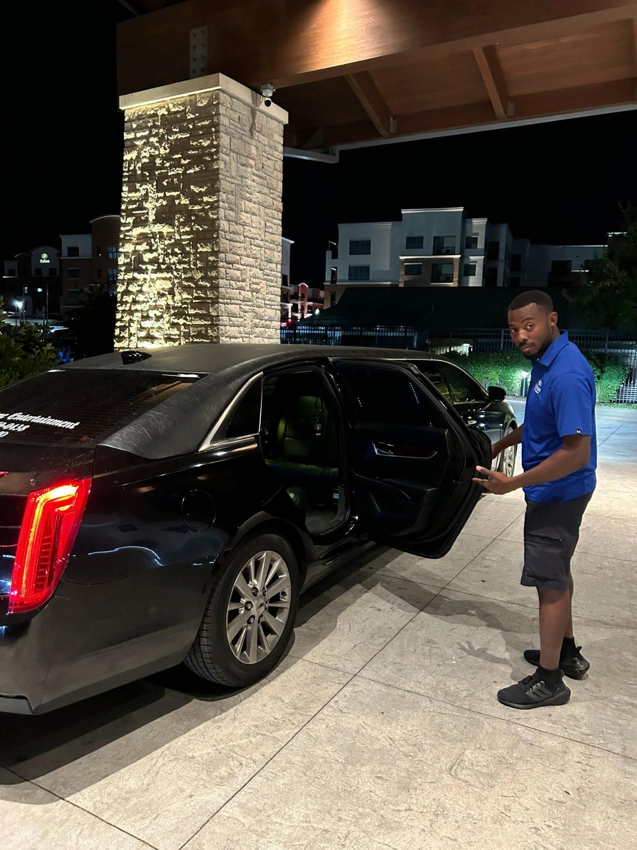 Man in blue shirt opens door of black sedan at night, near a building.
