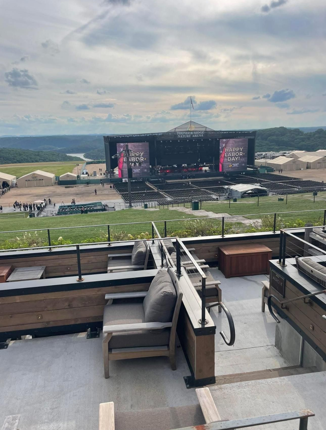 Outdoor concert stage on a hilltop. Empty seating area in foreground. Cloudy sky above.