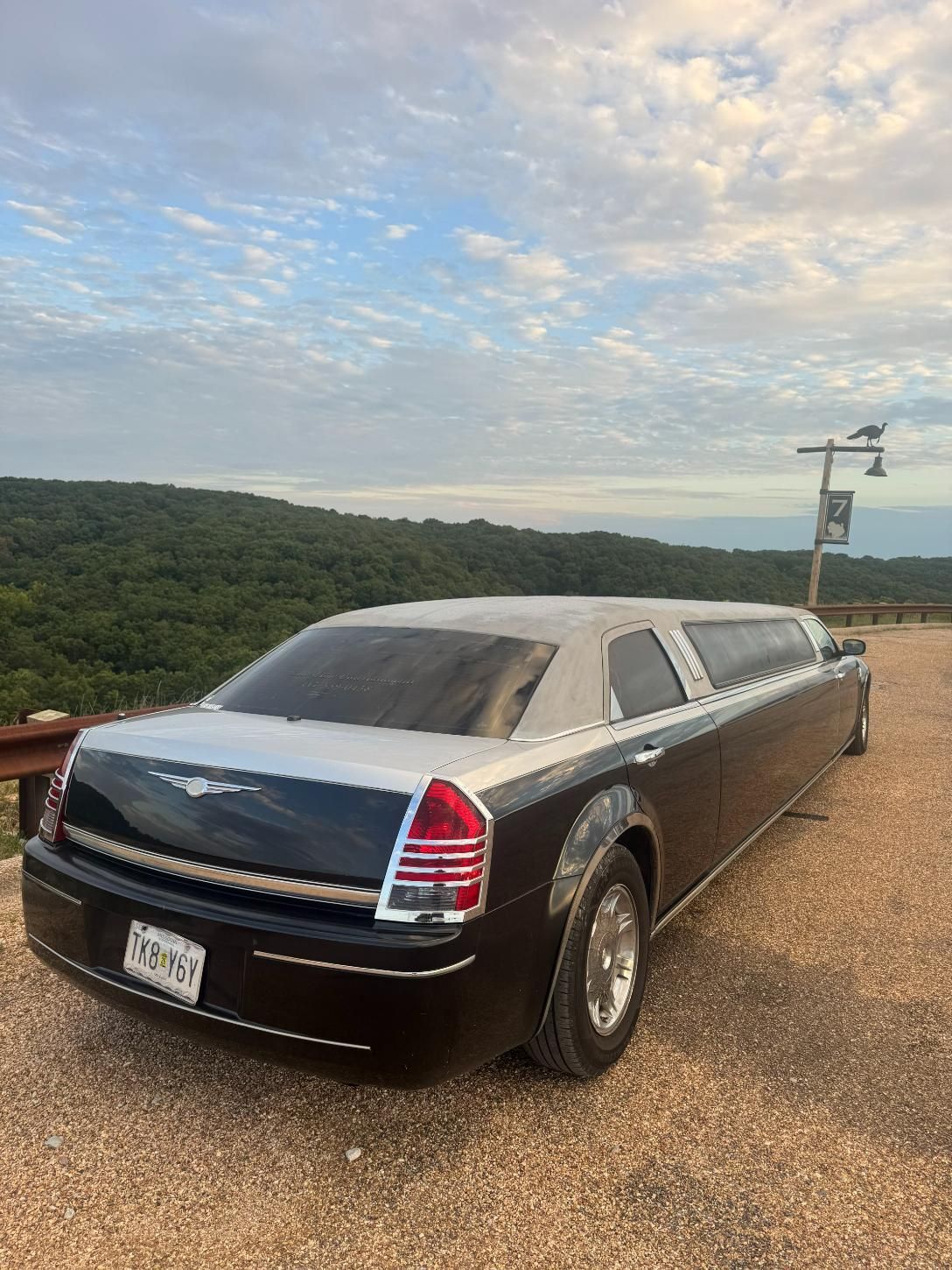 Black limousine on a rocky road overlooking a tree-covered hillside under a partly cloudy sky.