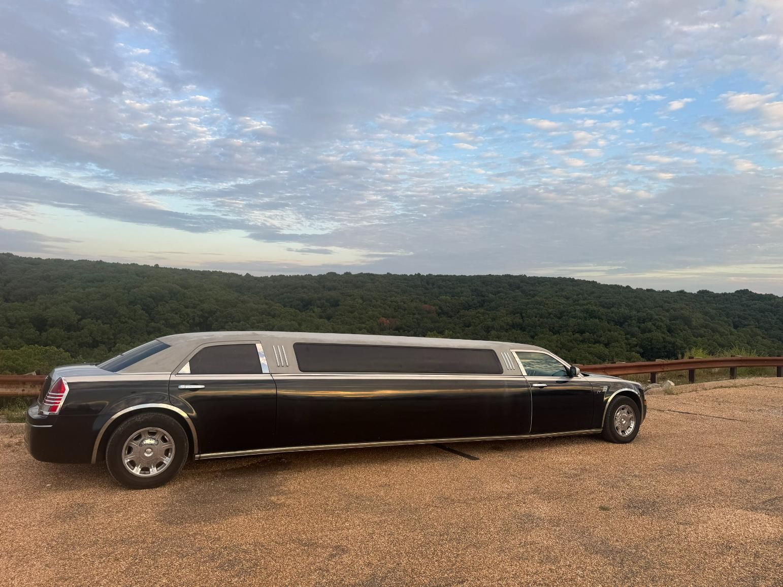 Black limousine parked on gravel road with a mountain backdrop and cloudy sky.
