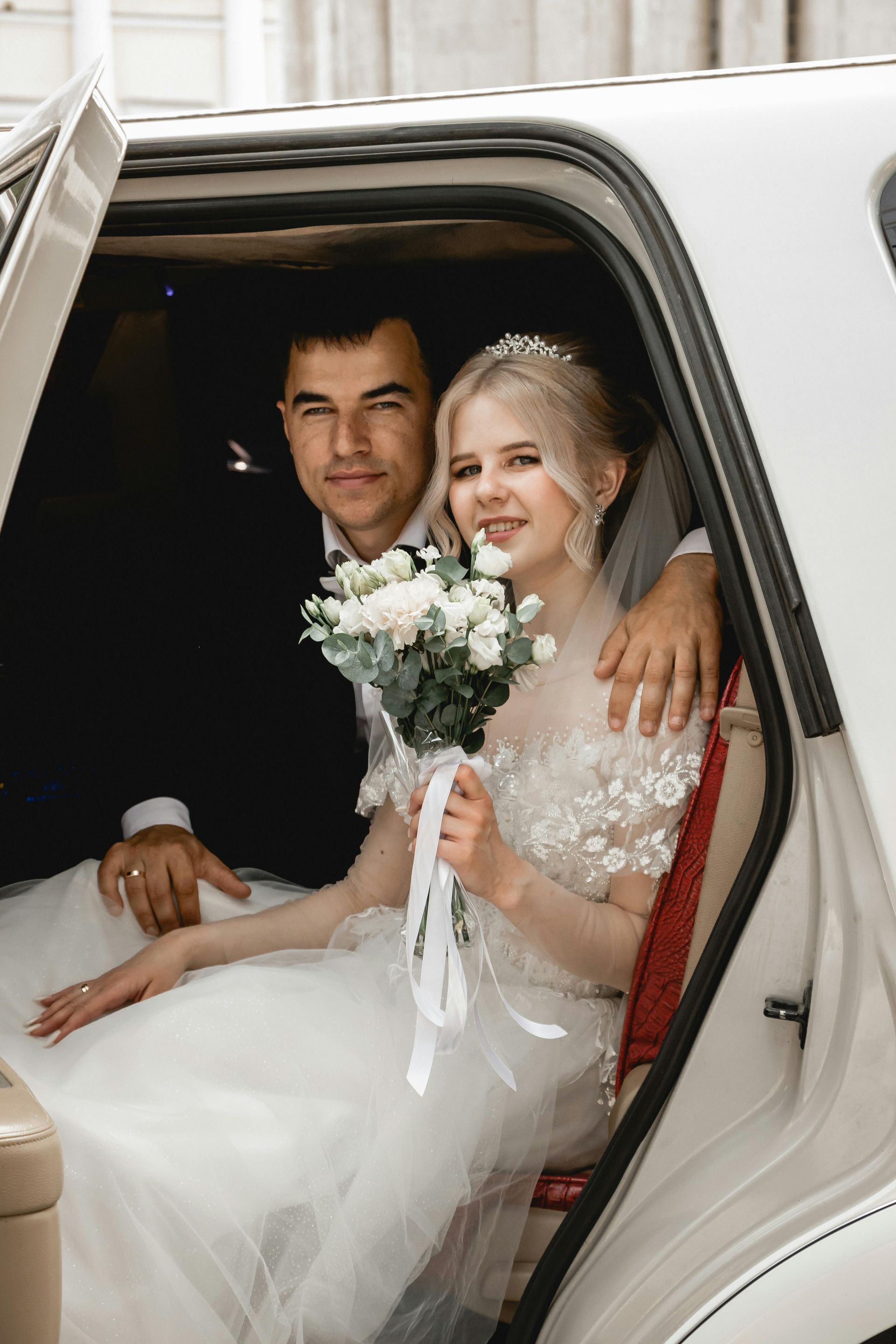 Newlyweds in a white car. The bride holds flowers, and the groom has his arm around her.