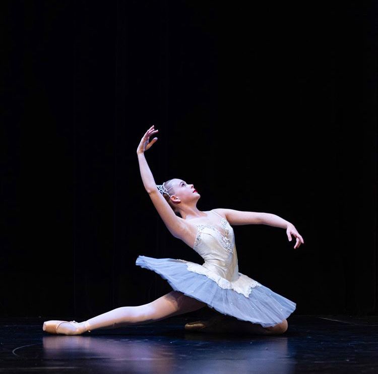 A ballerina in a white tutu is kneeling on the floor on a stage.