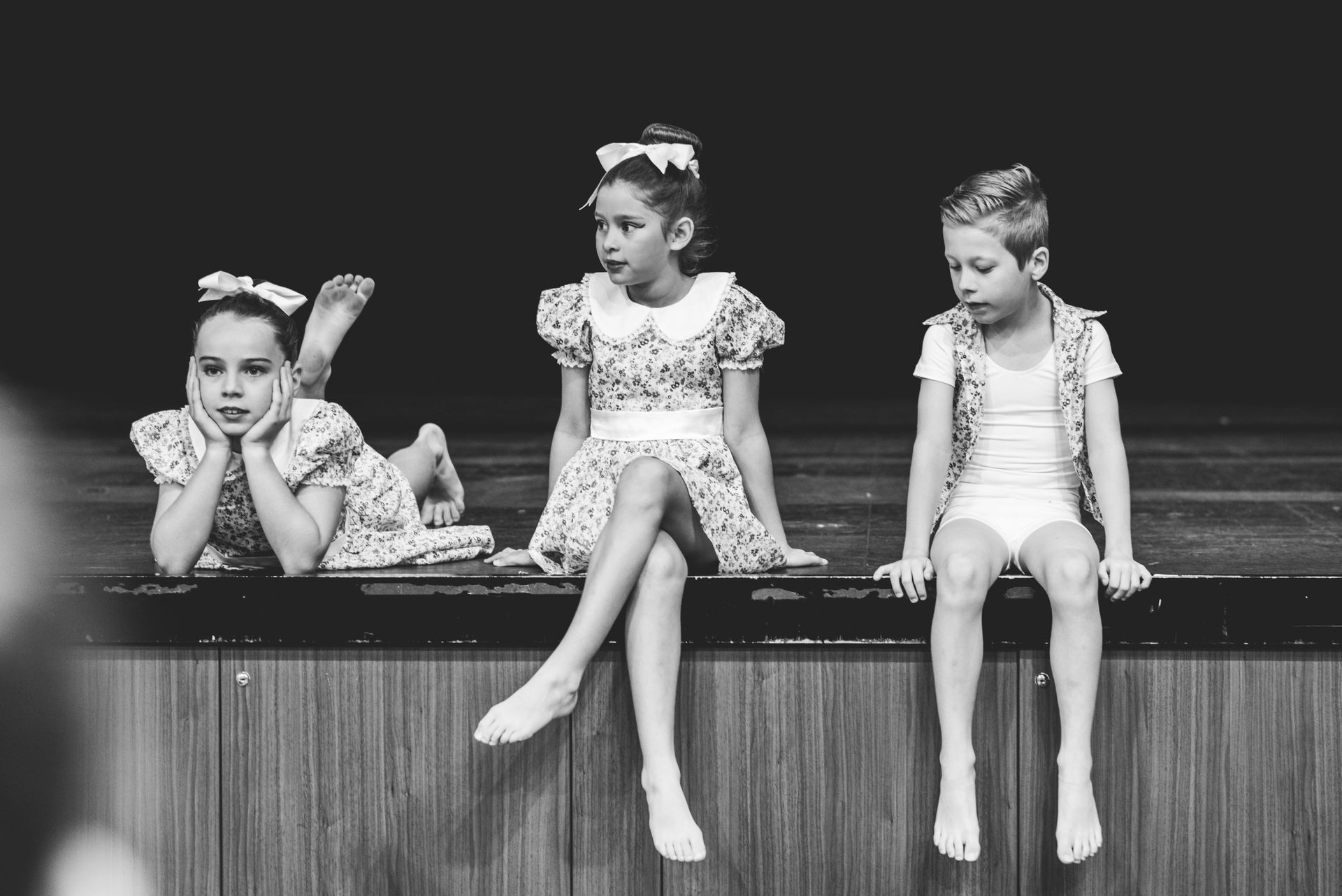 Three young girls are sitting on a wooden railing on a stage.