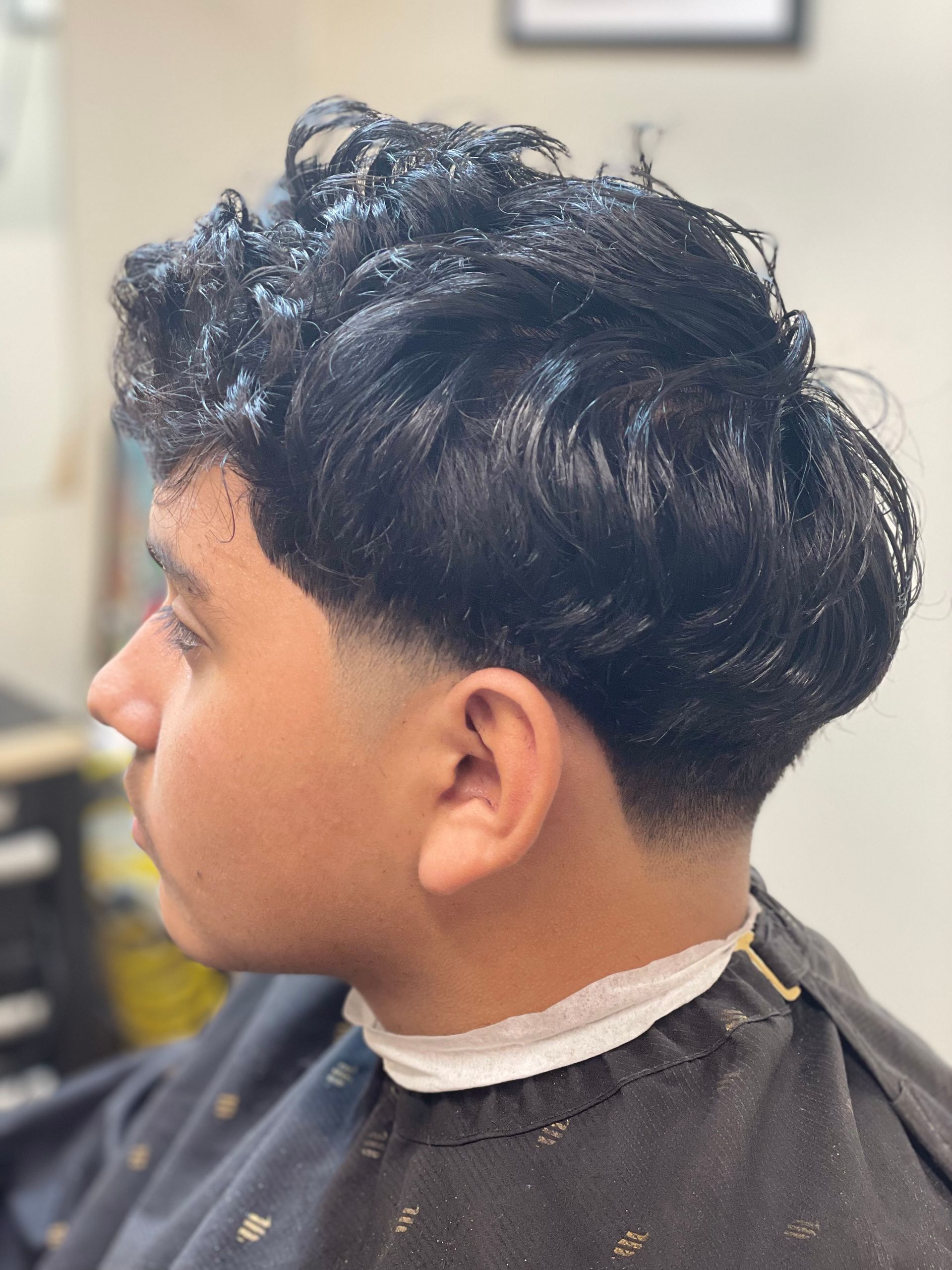 Man with a dark, wavy hairstyle, side profile with a faded haircut and a barber's cape.