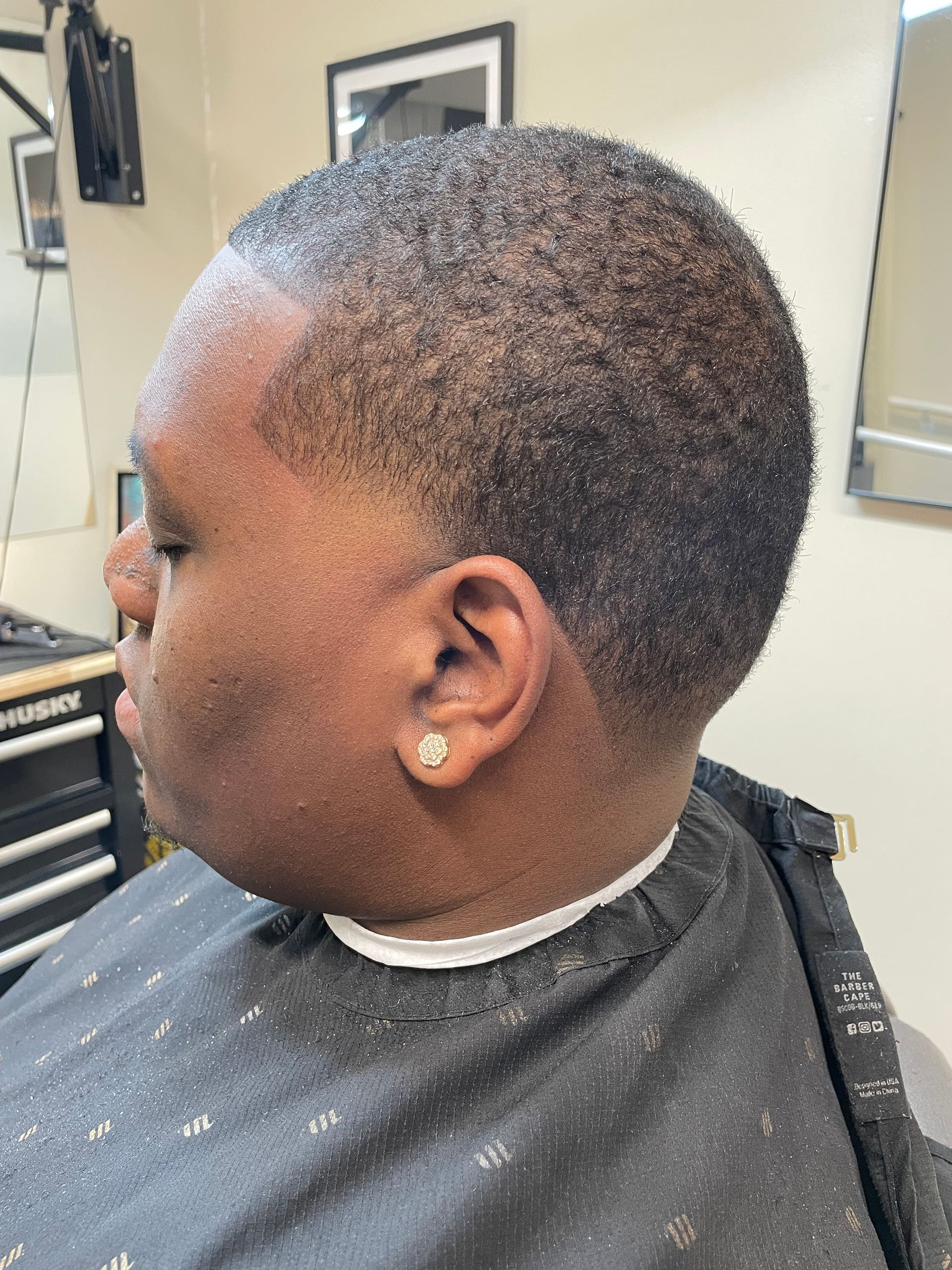 Person with short haircut, in a barber's chair, side view. Dark skin, wearing a barber's cape.