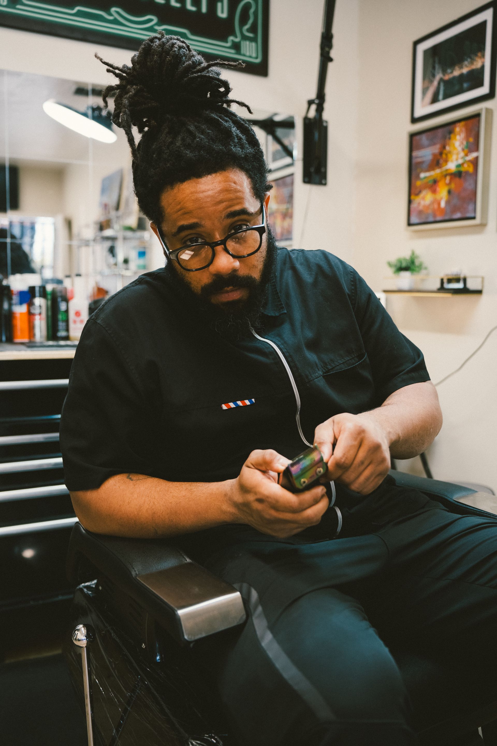 Man in barbershop, holding object, looking at camera. Wearing glasses, black shirt, hair in bun.