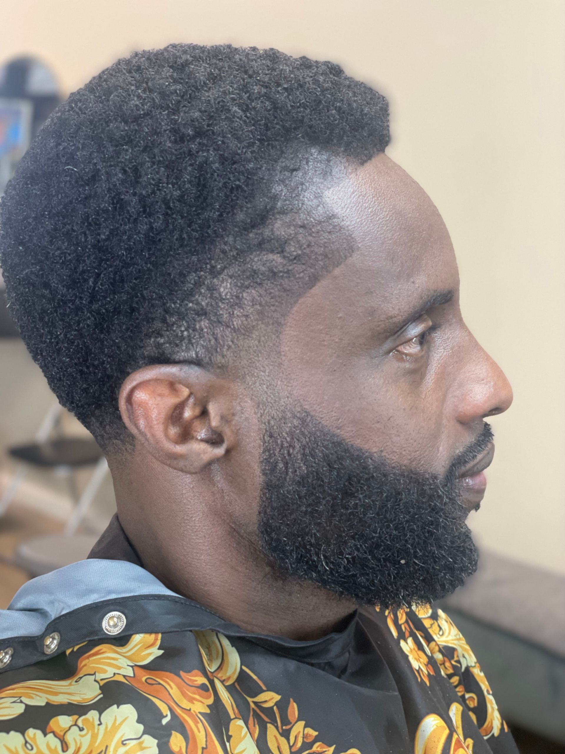 Man with dark skin, side profile. Short, curly hair, trimmed beard. Wearing a patterned top.