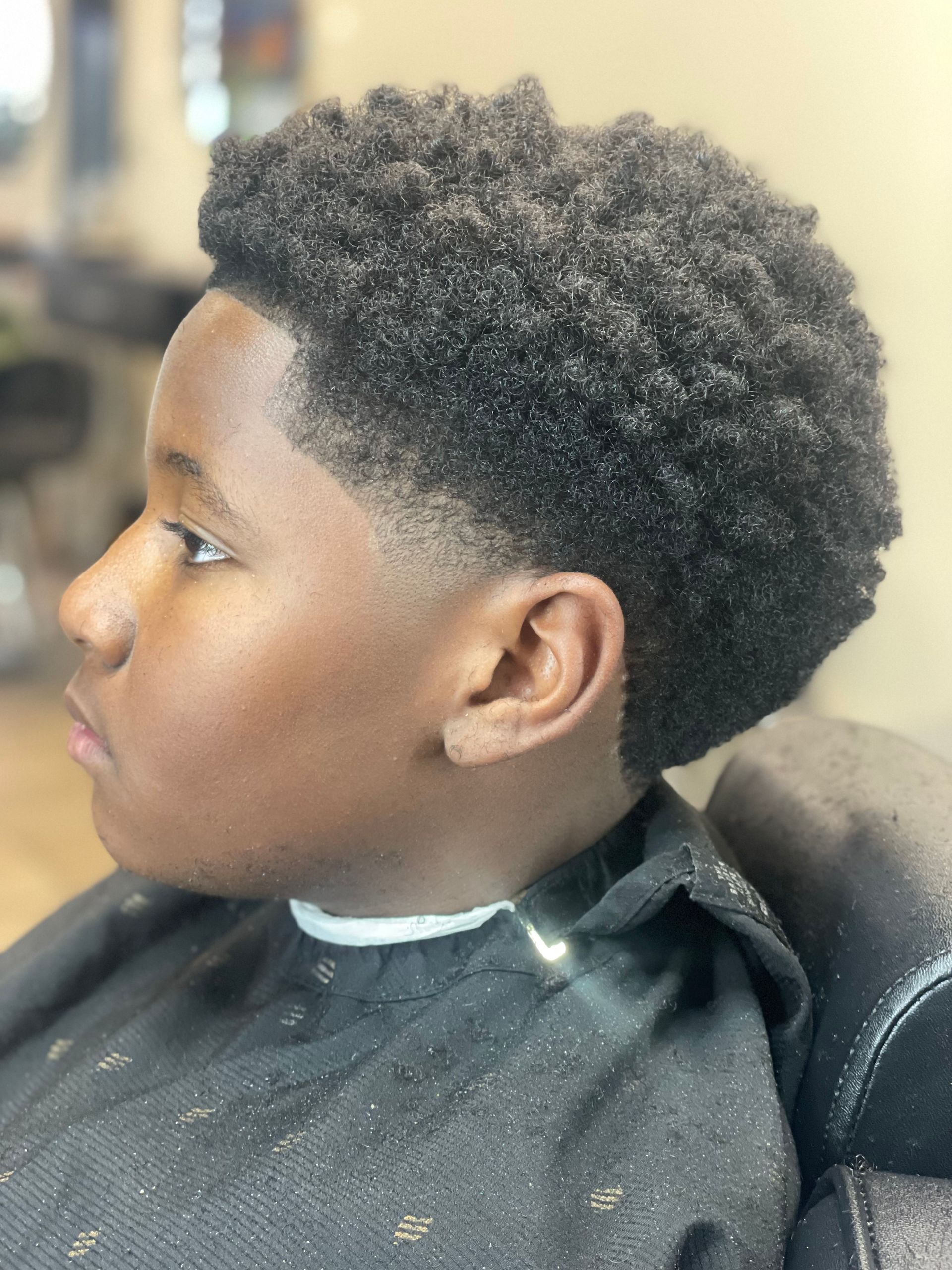 Boy with afro hairstyle, fade haircut on the sides, black cape in a barbershop setting.