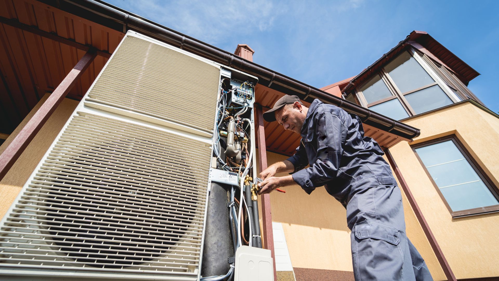 A technician repairs an outdoor air conditioning unit on the side of a house. Bright blue sky.