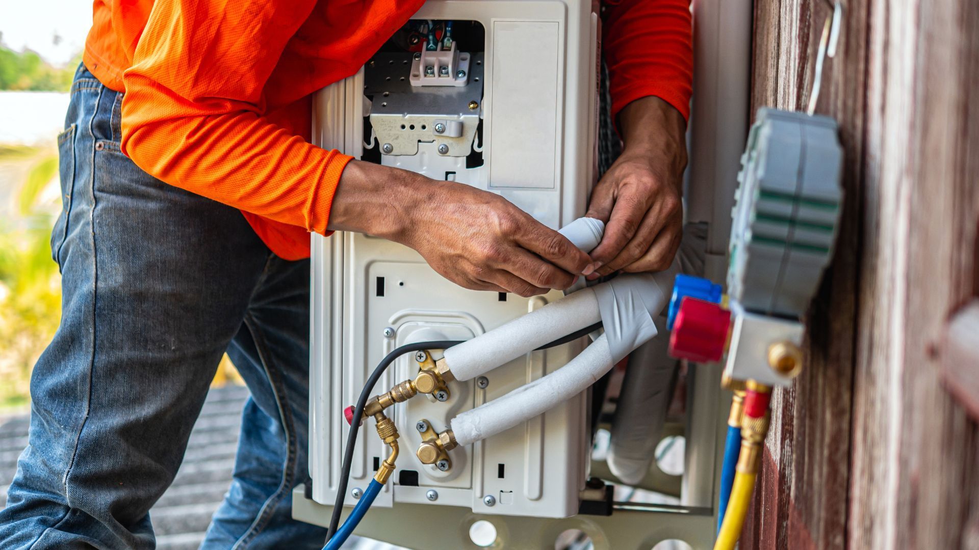 A repairman in an orange shirt and jeans works on an AC unit outside. He is connecting a white tube.