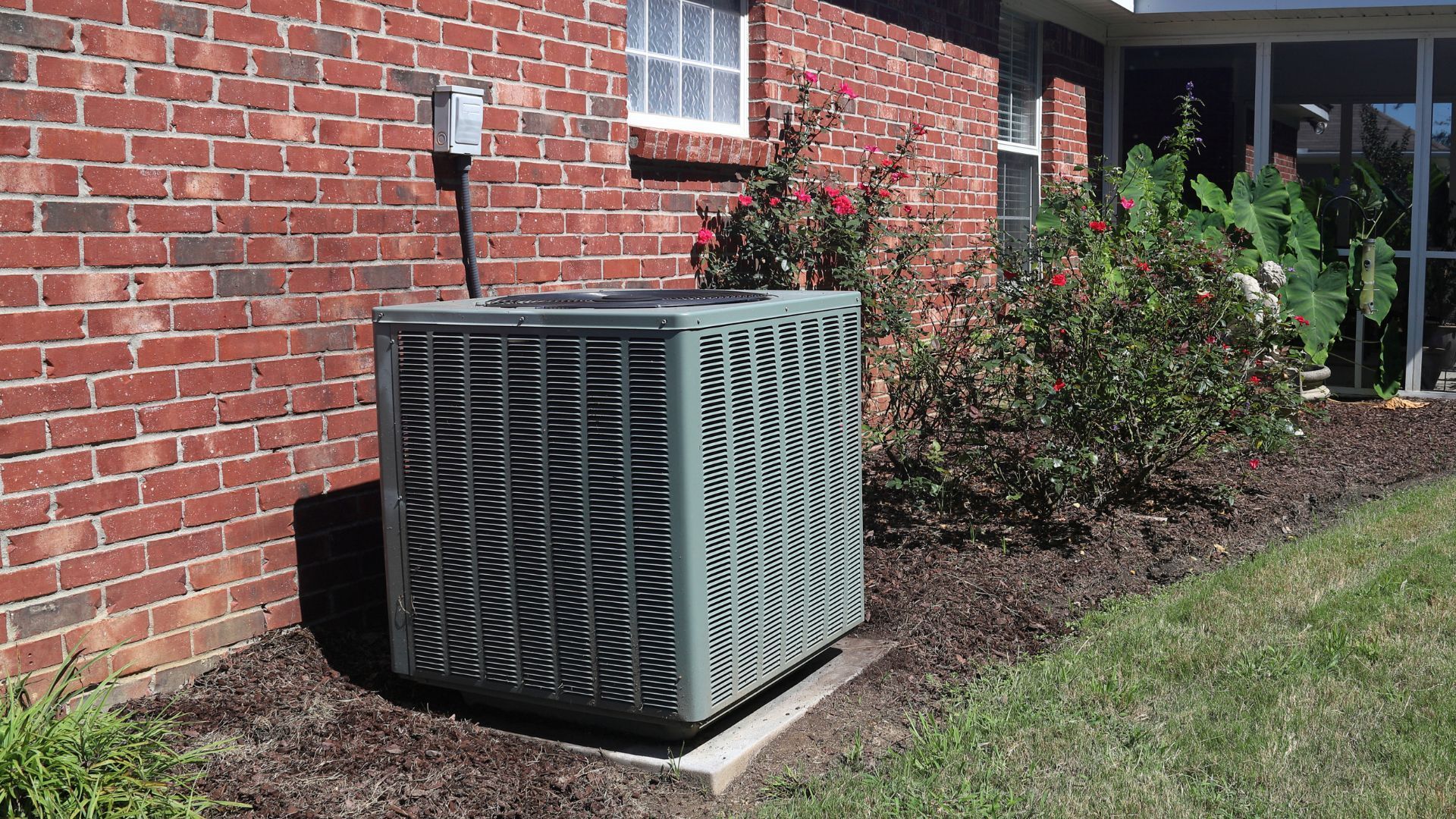 An outdoor air conditioning unit next to a red brick wall and landscaping.