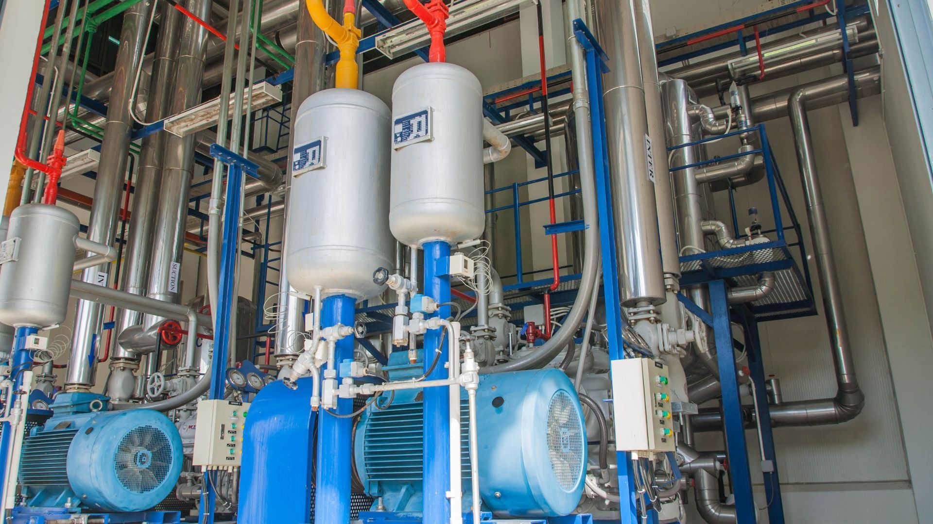 Industrial equipment with blue pumps, silver tanks, and colorful pipes in a factory setting.