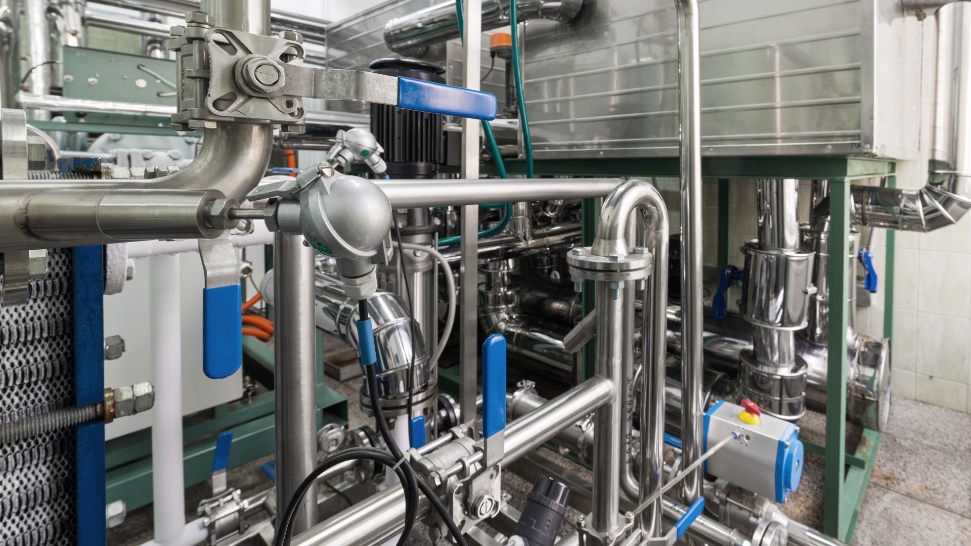 Complex network of industrial pipes, valves, and equipment in a factory setting. Shiny metal against a background of white and green.