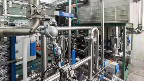 Complex network of industrial pipes, valves, and equipment in a factory setting. Shiny metal against a background of white and green.
