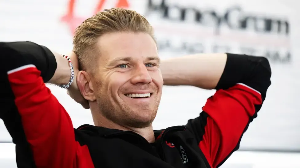 A man in a red and black shirt is smiling with his hands behind his head.