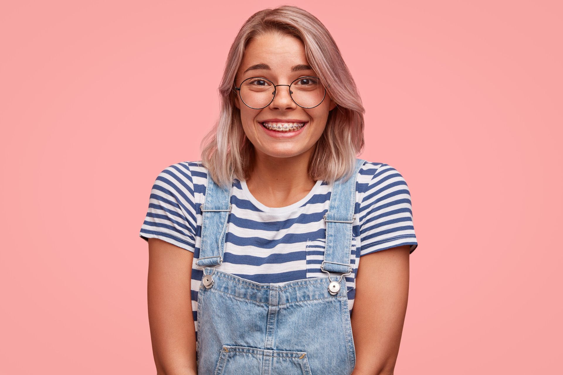 A young woman wearing glasses and overalls is smiling.