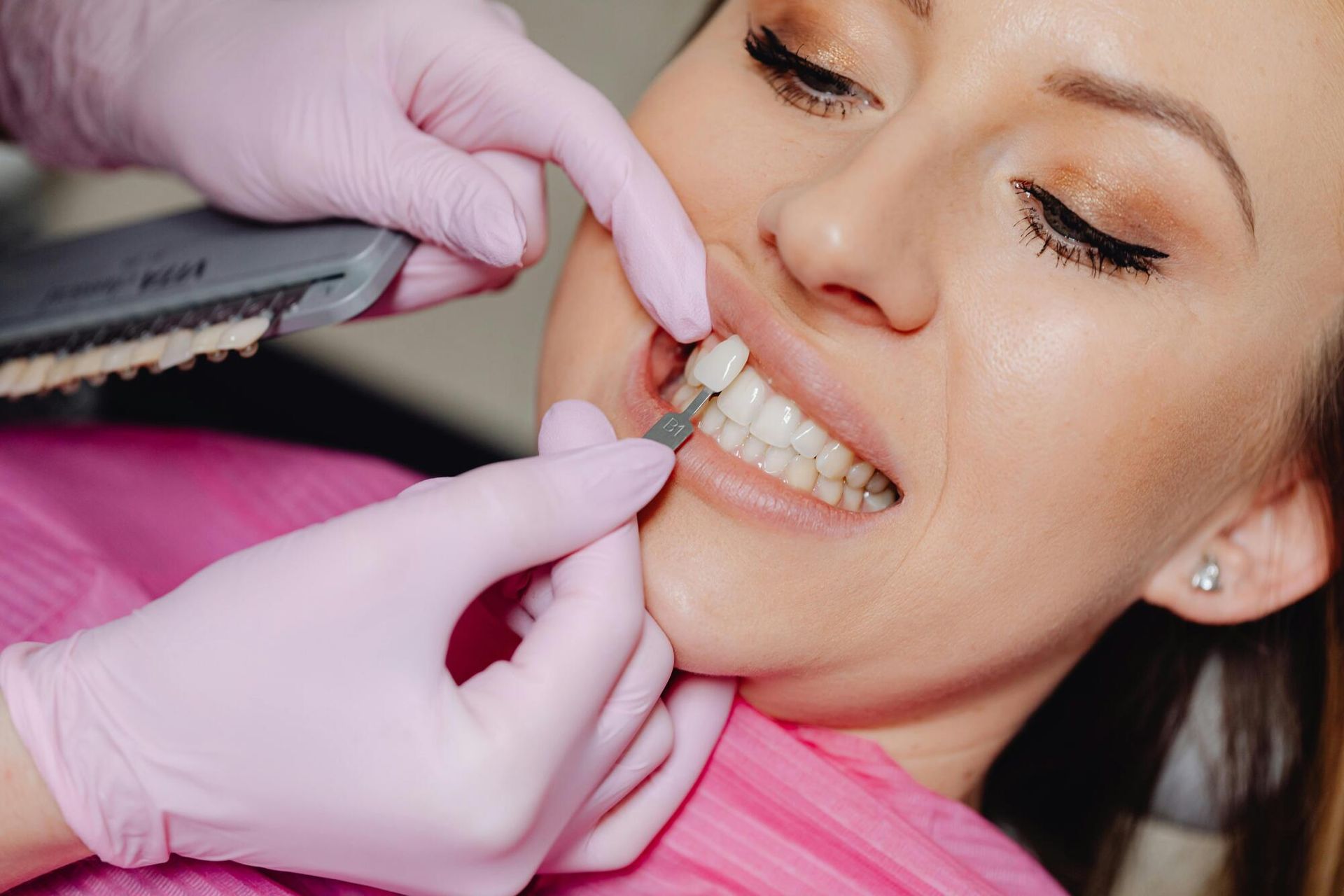 Dentist matching patient's teeth color with shade guide. Pink gloves and bib, dental setting.