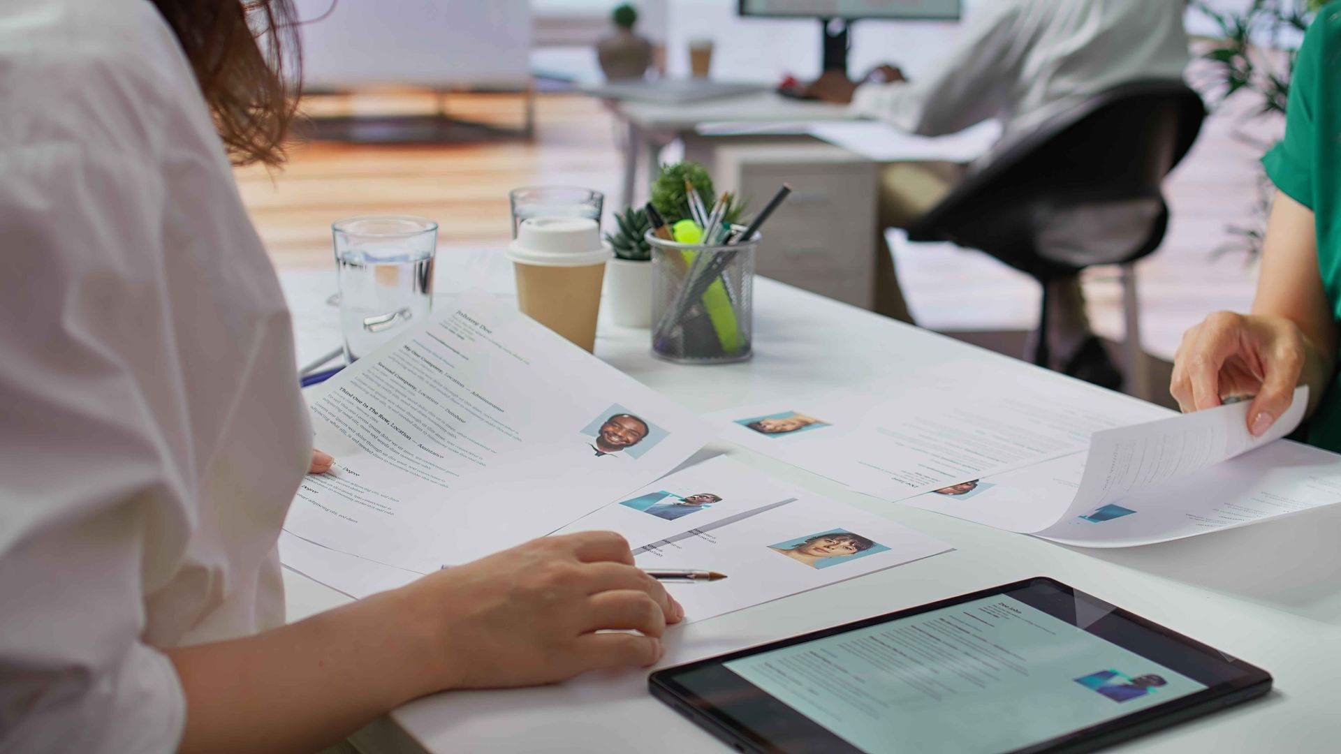 People reviewing documents and a tablet at a desk, with a person sitting at a computer in the background.
