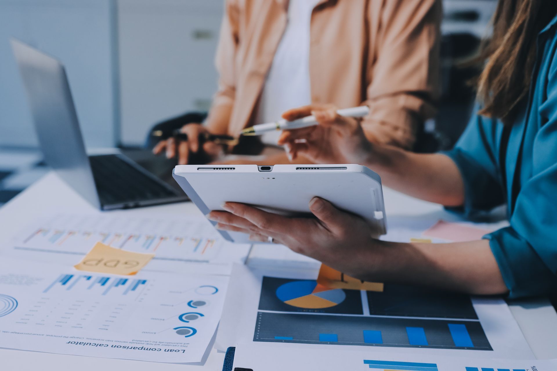 Two people review financial charts and data on a tablet and laptop, collaborating in an office.