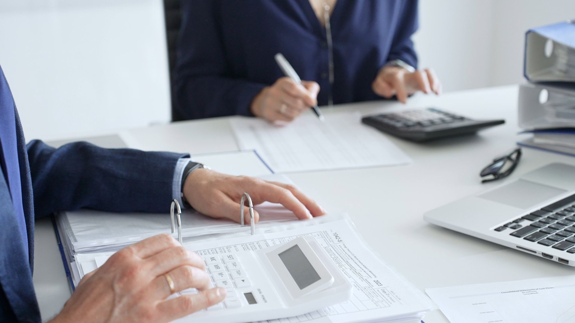 Two people in business suits working with papers, calculators, and a laptop.