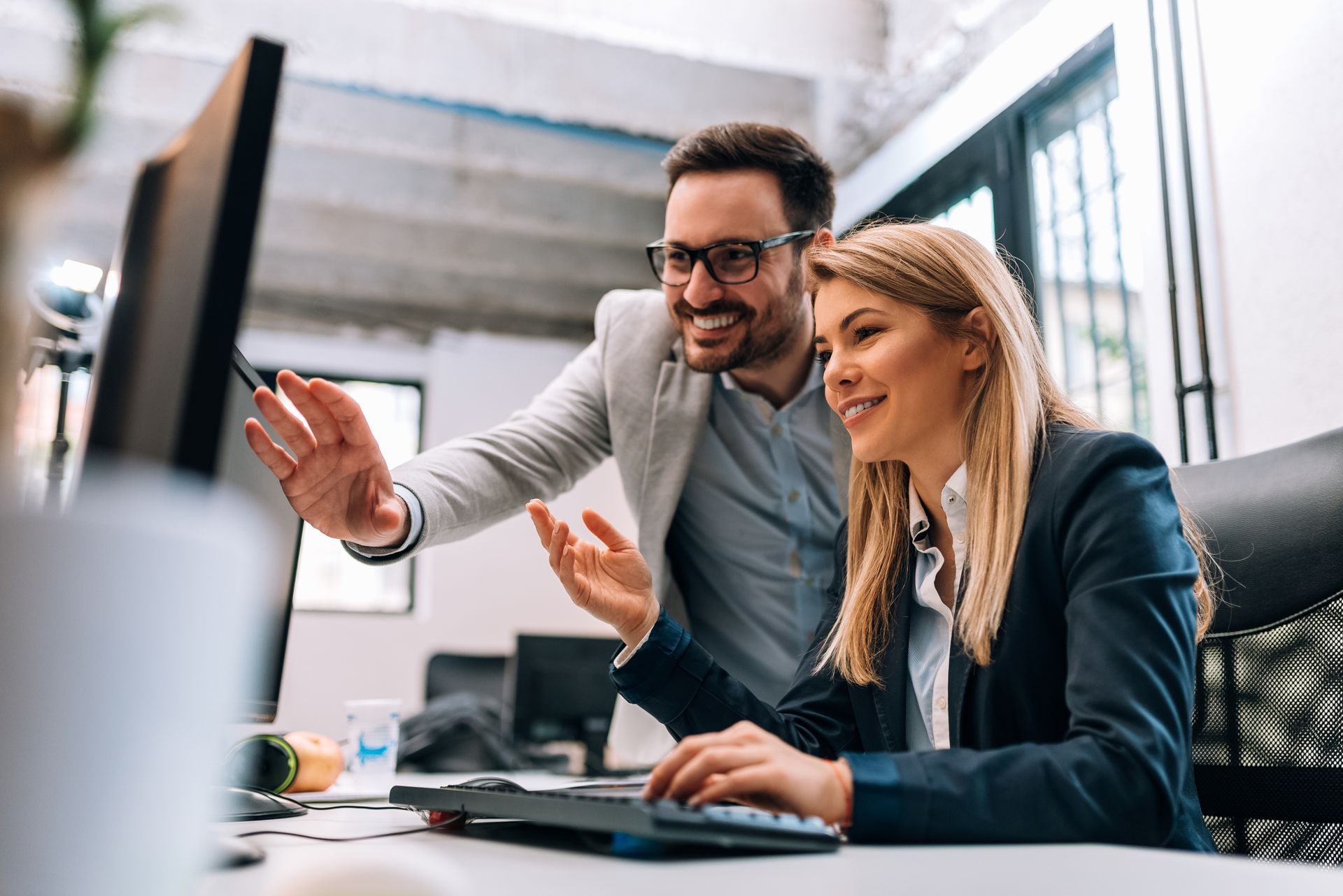 Man points at computer screen, smiling at woman typing. Office setting.