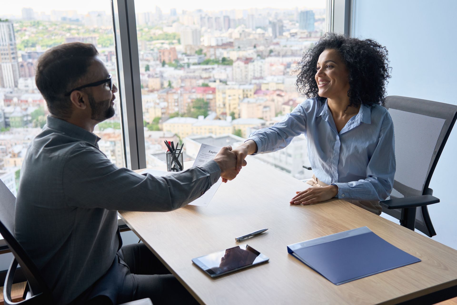 Two people shaking hands at a desk in an office overlooking a city.