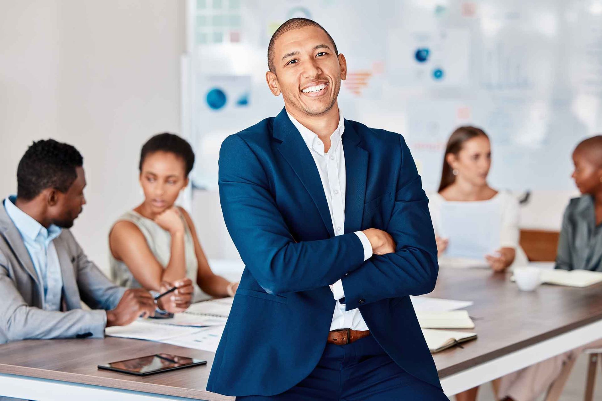 Man in blue suit smiles, arms crossed, in a meeting with colleagues.