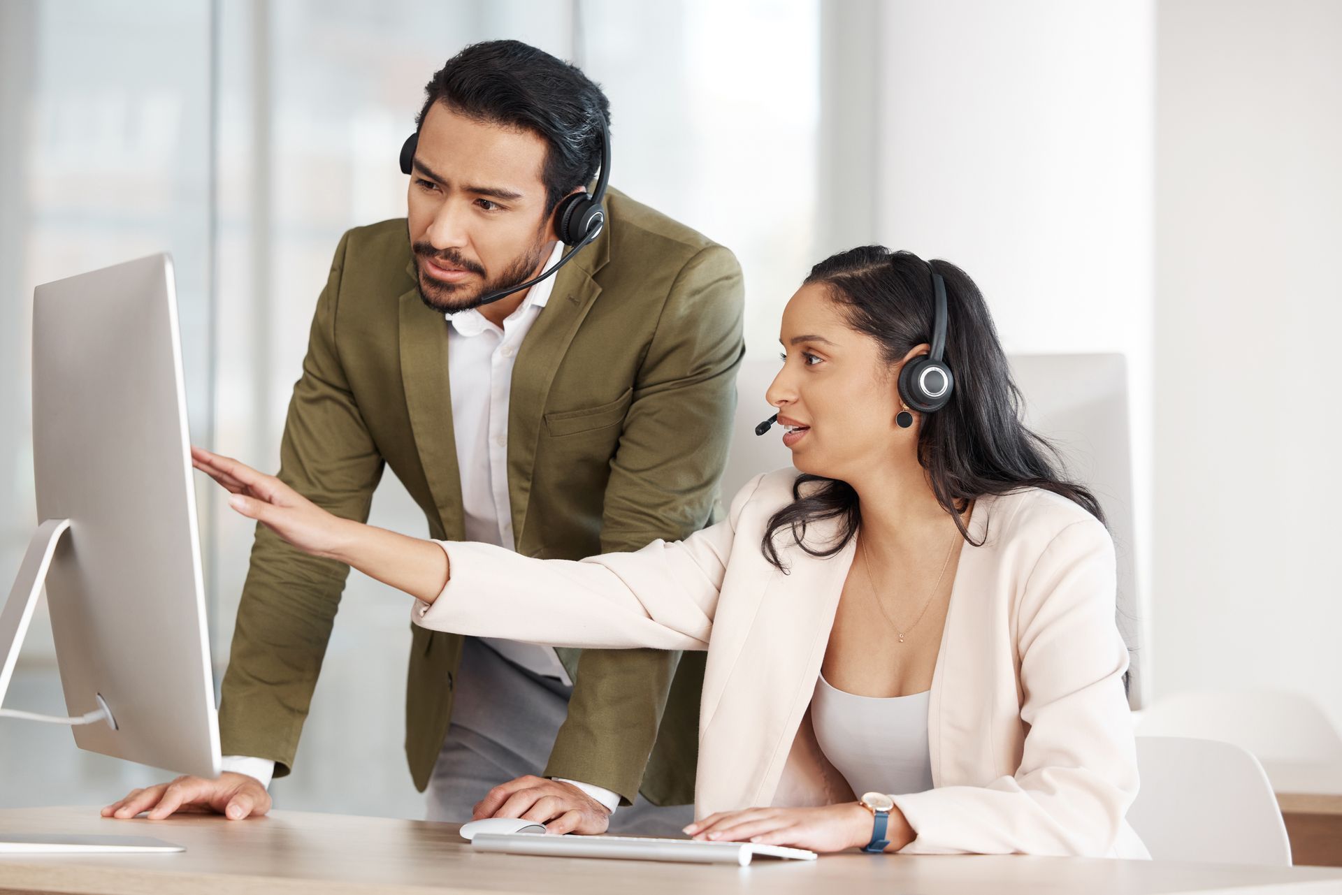 Two people wearing headsets look at a computer. One points at the screen while the other looks on.