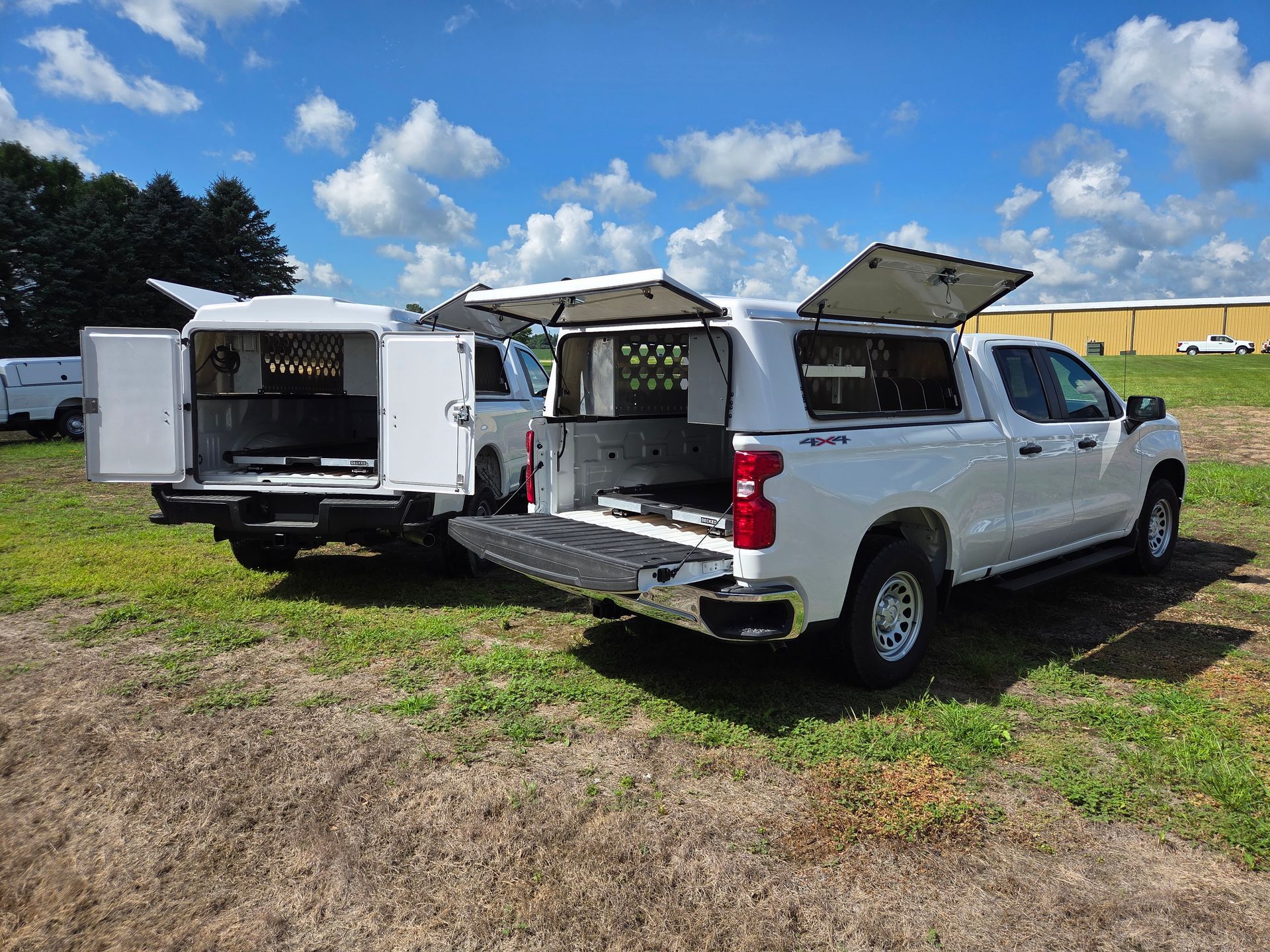 Two white pickup trucks with utility bed caps, parked in a field with open doors.