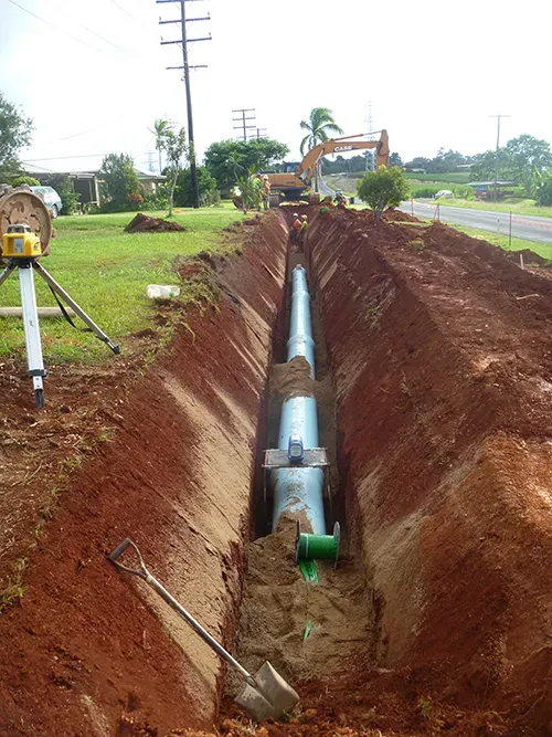 Construction site: a trench with large pipes, excavator, shovel, and leveler on grass by a road — Far North Plumbing in Mourilyan, QLD