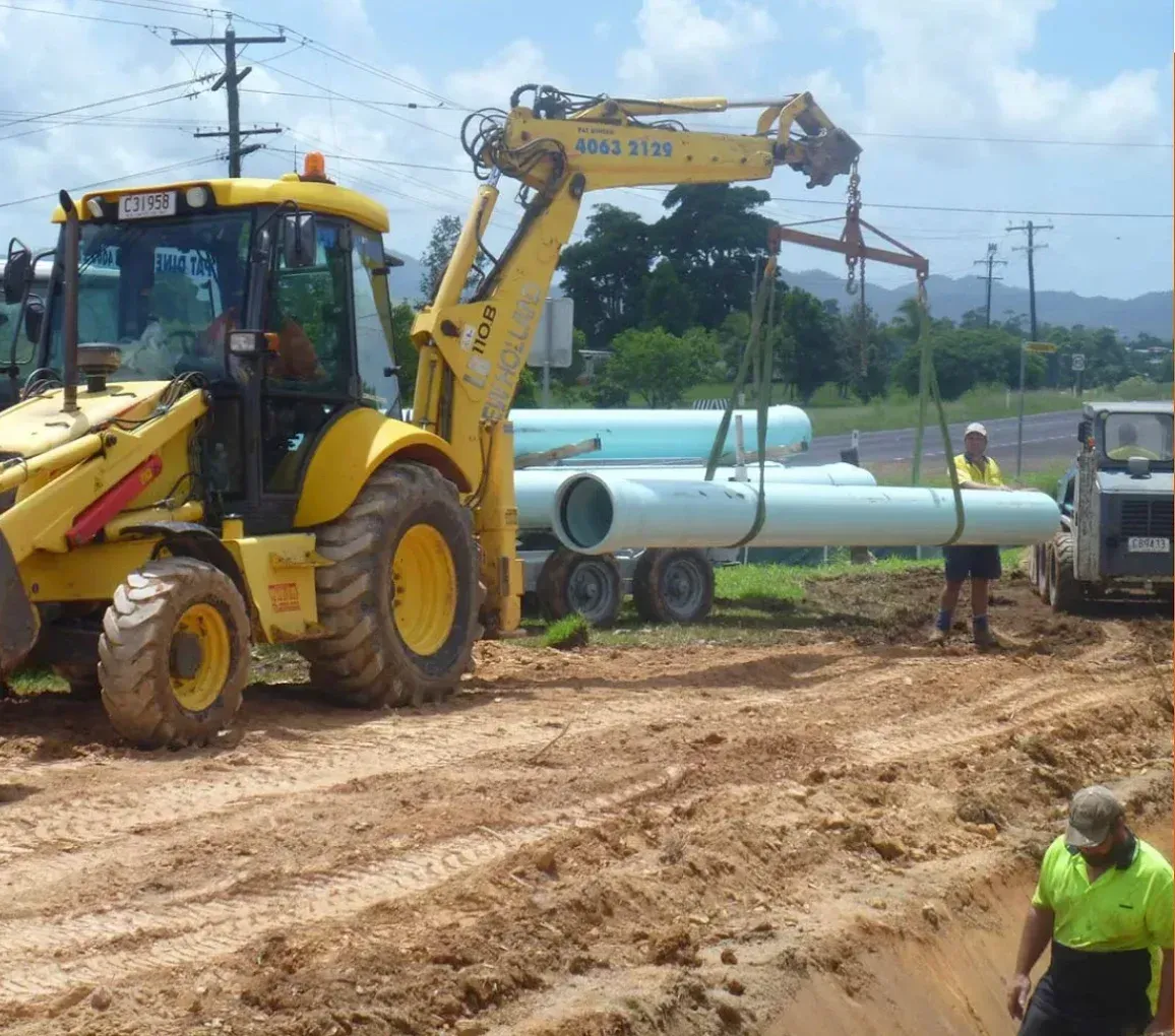 Yellow backhoe lifting pipes from a trailer at a construction site. Two workers present, sunny day — Far North Plumbing in Mourilyan, QLD