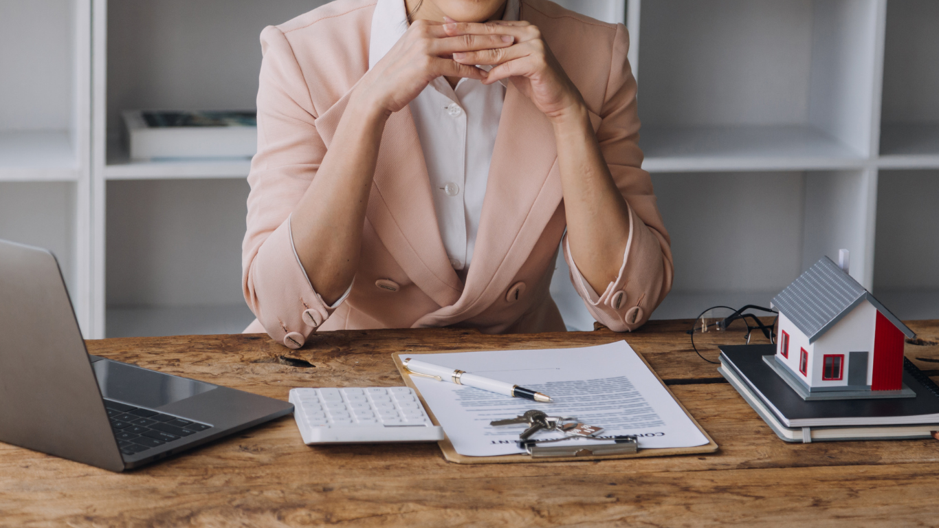 Woman in pink blazer at a desk, hands on chin. Documents, keys, house model, and laptop.