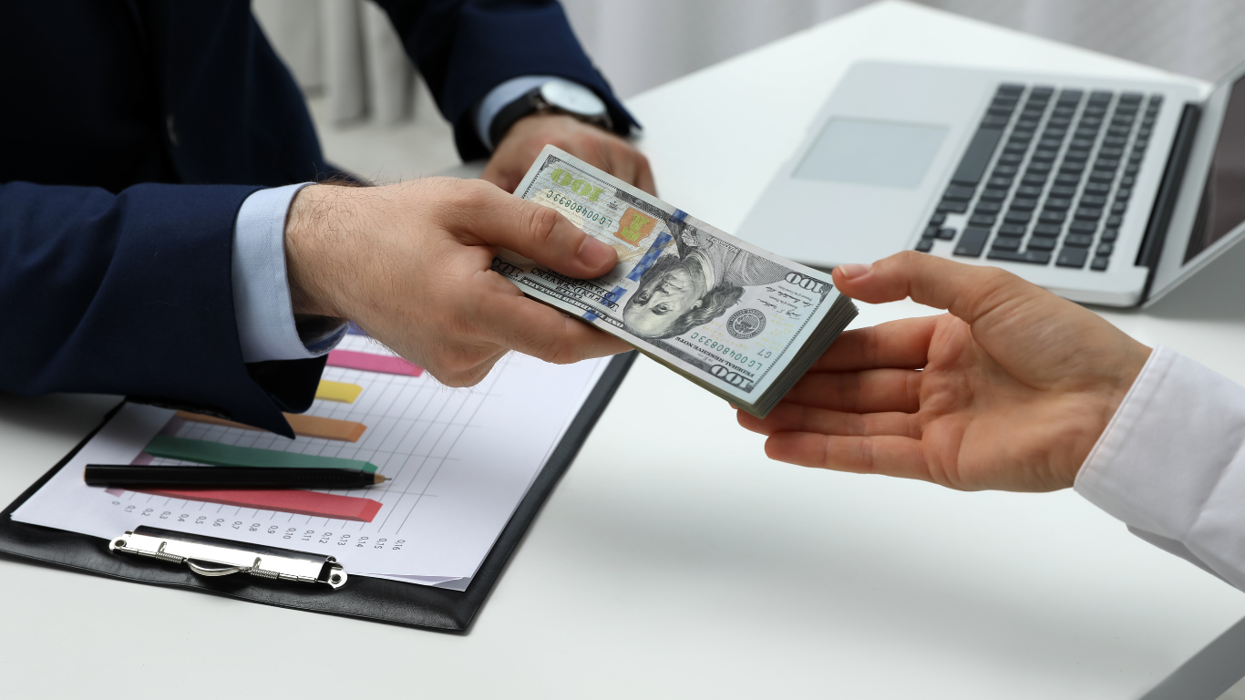 Person in suit hands a stack of US dollar bills to another person at a desk with a laptop and documents.