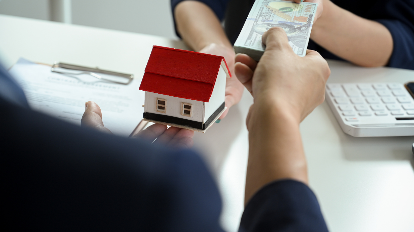 Person handing cash to another person next to a toy house; transaction.