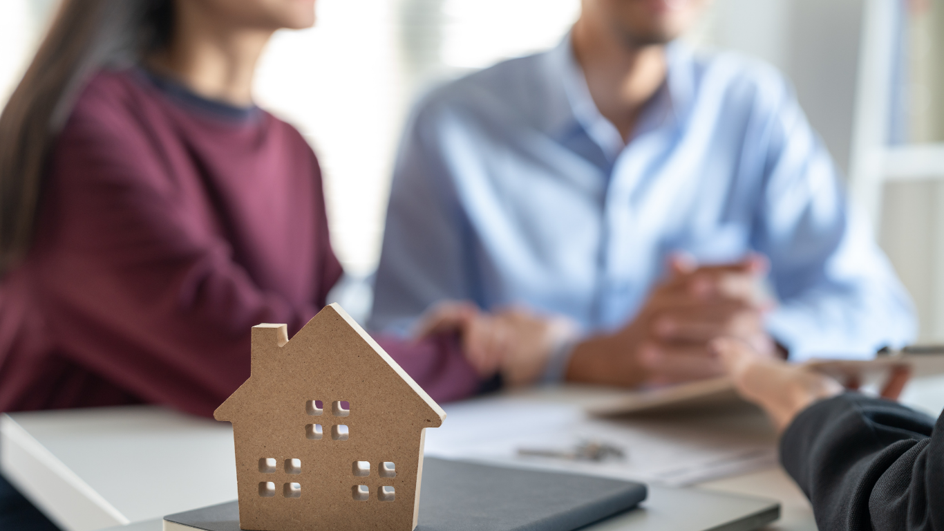 Couple reviewing paperwork with realtor; wooden house model on table.