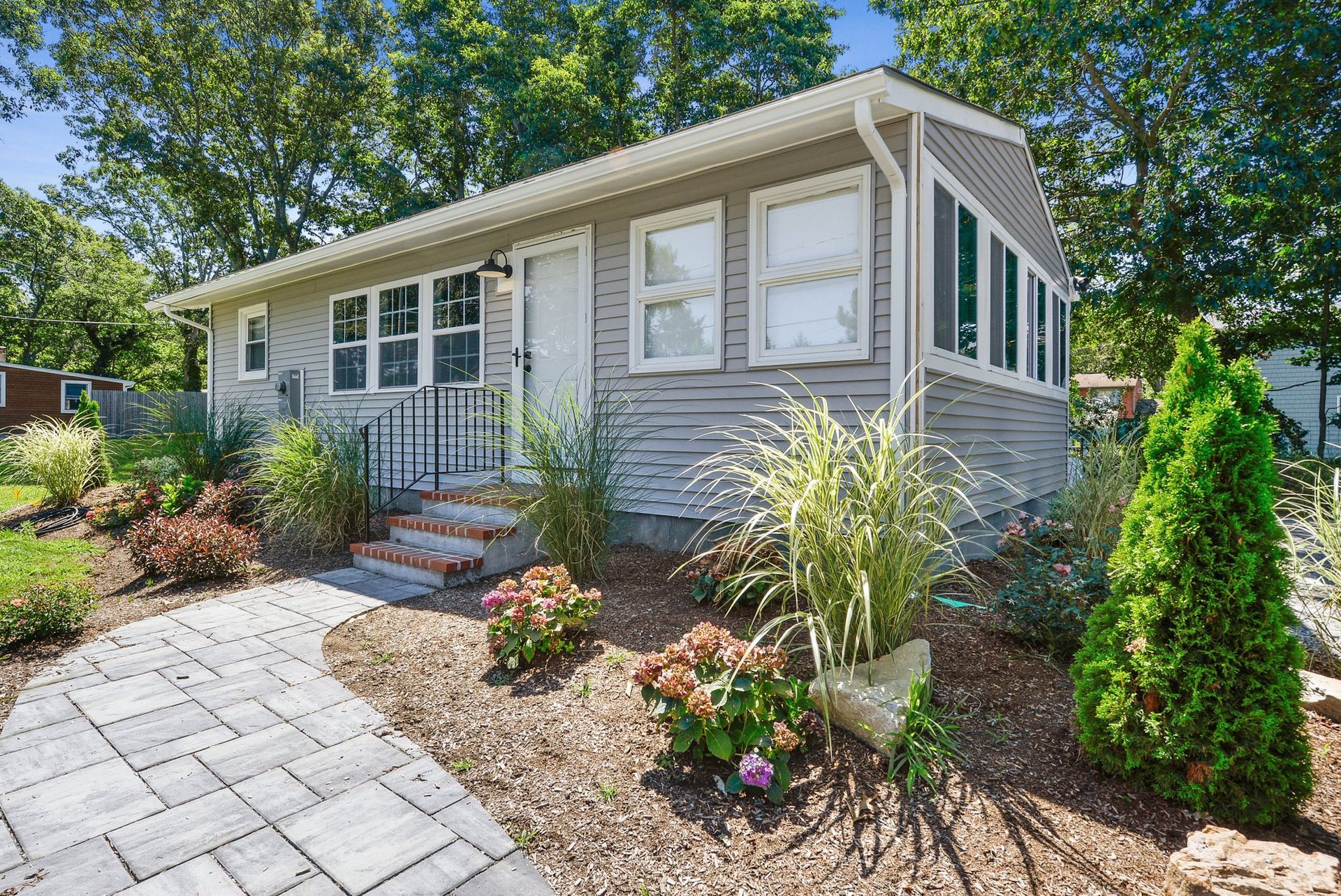 A small, gray-sided house with a paved walkway, front steps, and a landscaped yard with green bushes and ornamental grass.