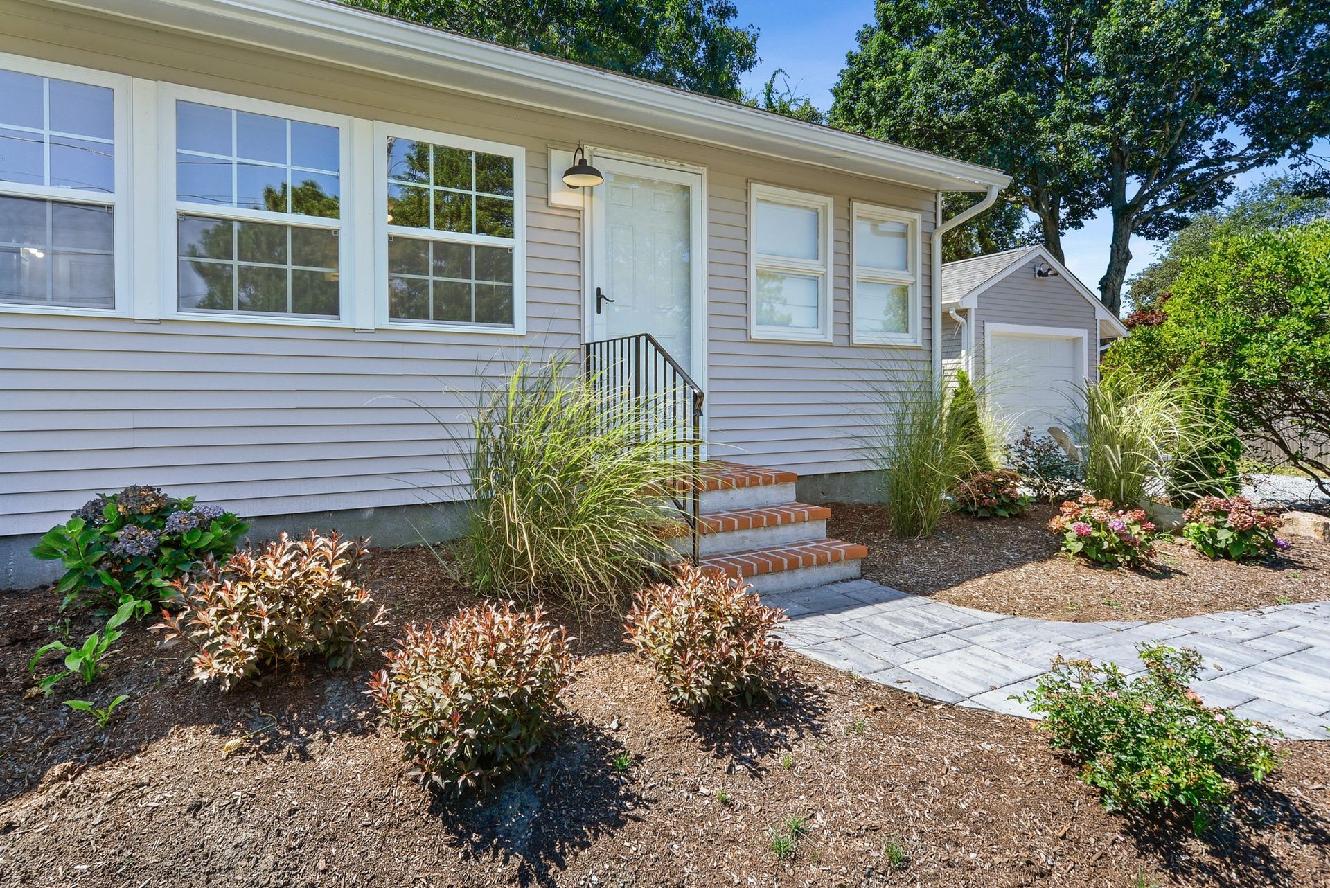 A light gray house with white trim, a small front porch, and a landscaped front yard with mulch and green shrubs.