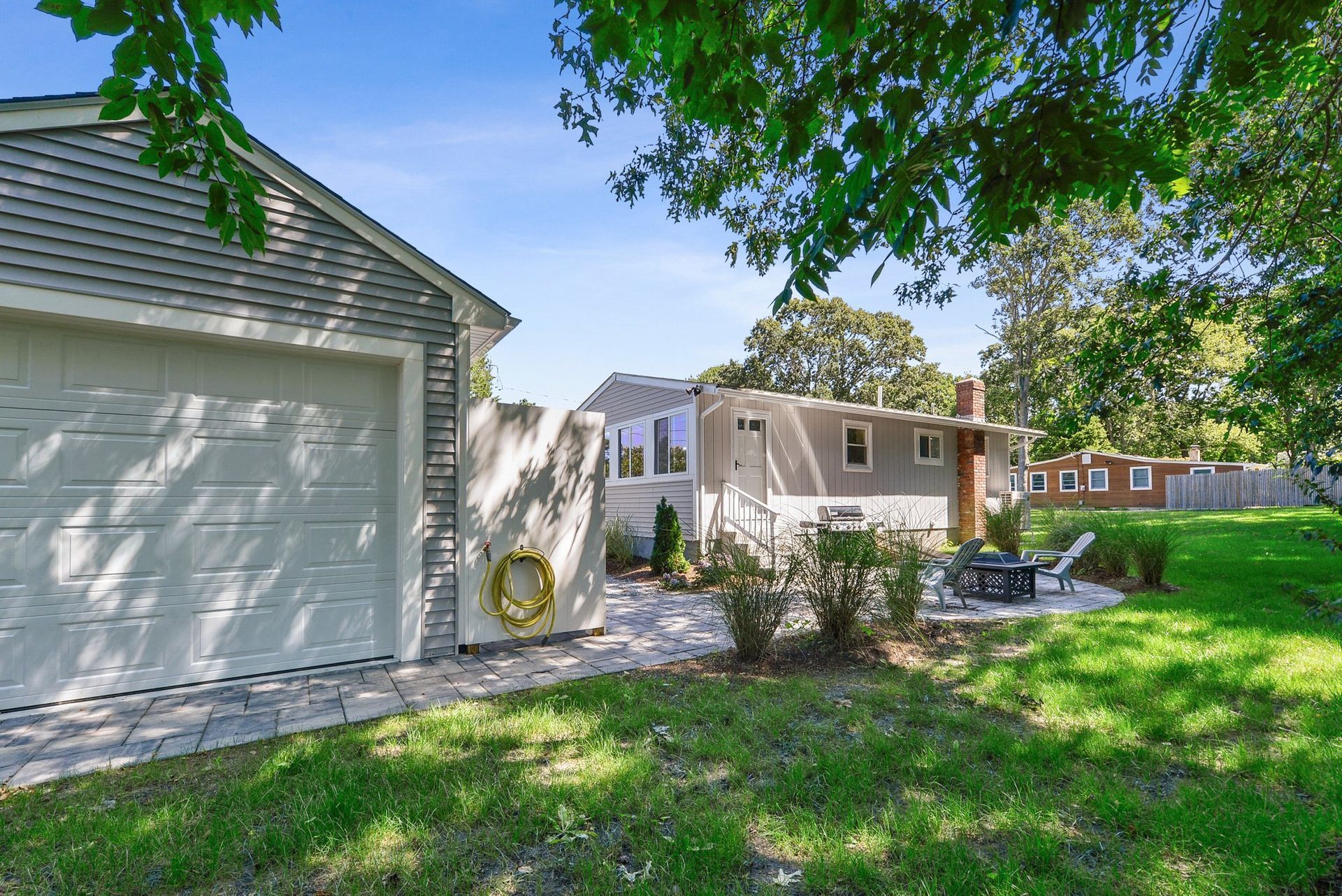 A suburban backyard with a gray detached garage, a white house with a patio, and a grassy lawn under a sunny blue sky.