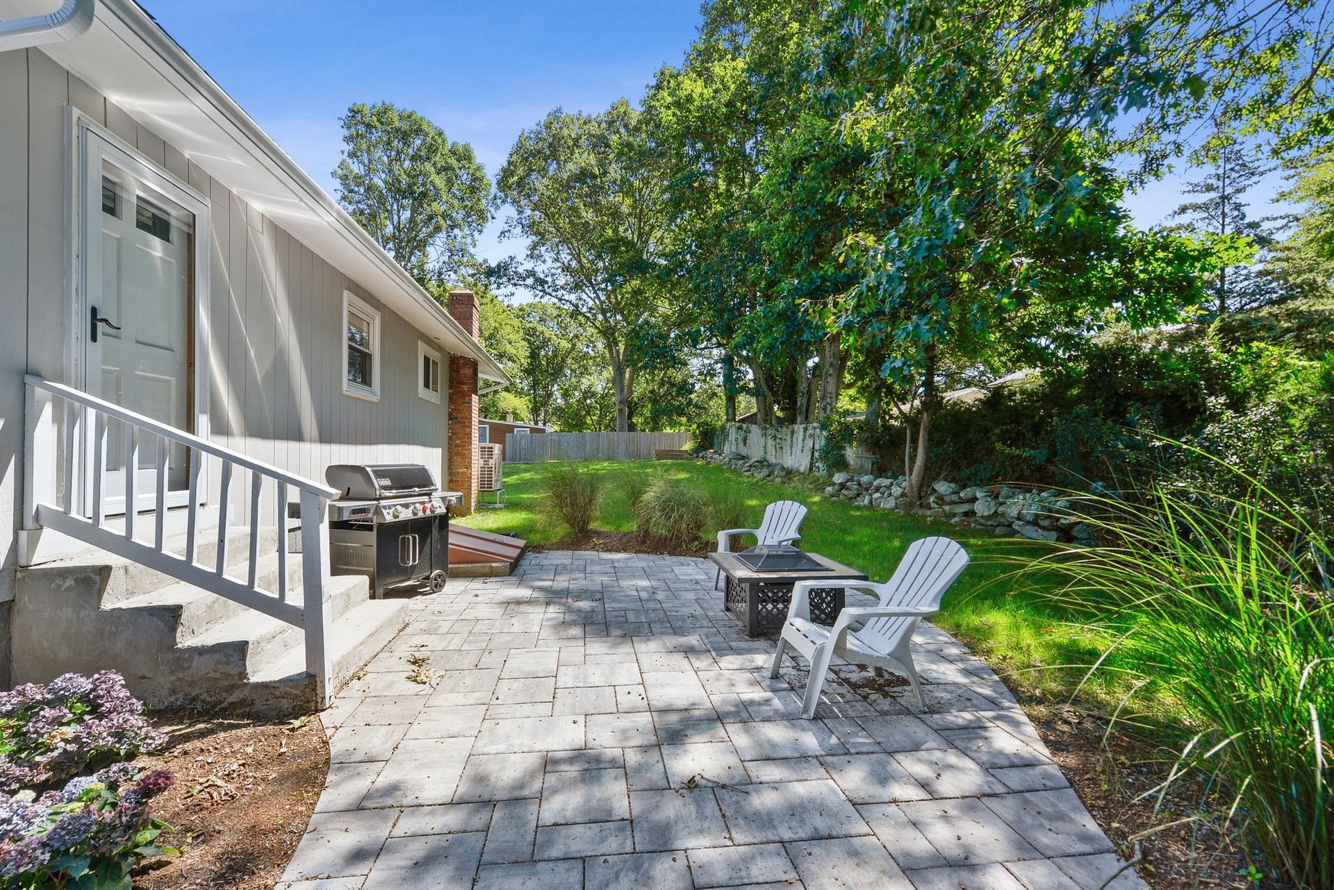 A gray house with a stone patio, outdoor grill, and two white chairs in a sunny, tree-filled backyard.