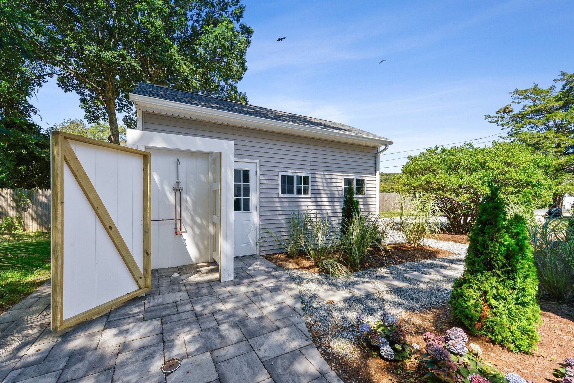 An outdoor shower enclosure next to a gray-sided building with a paved patio, trees, and landscaping under a blue sky.