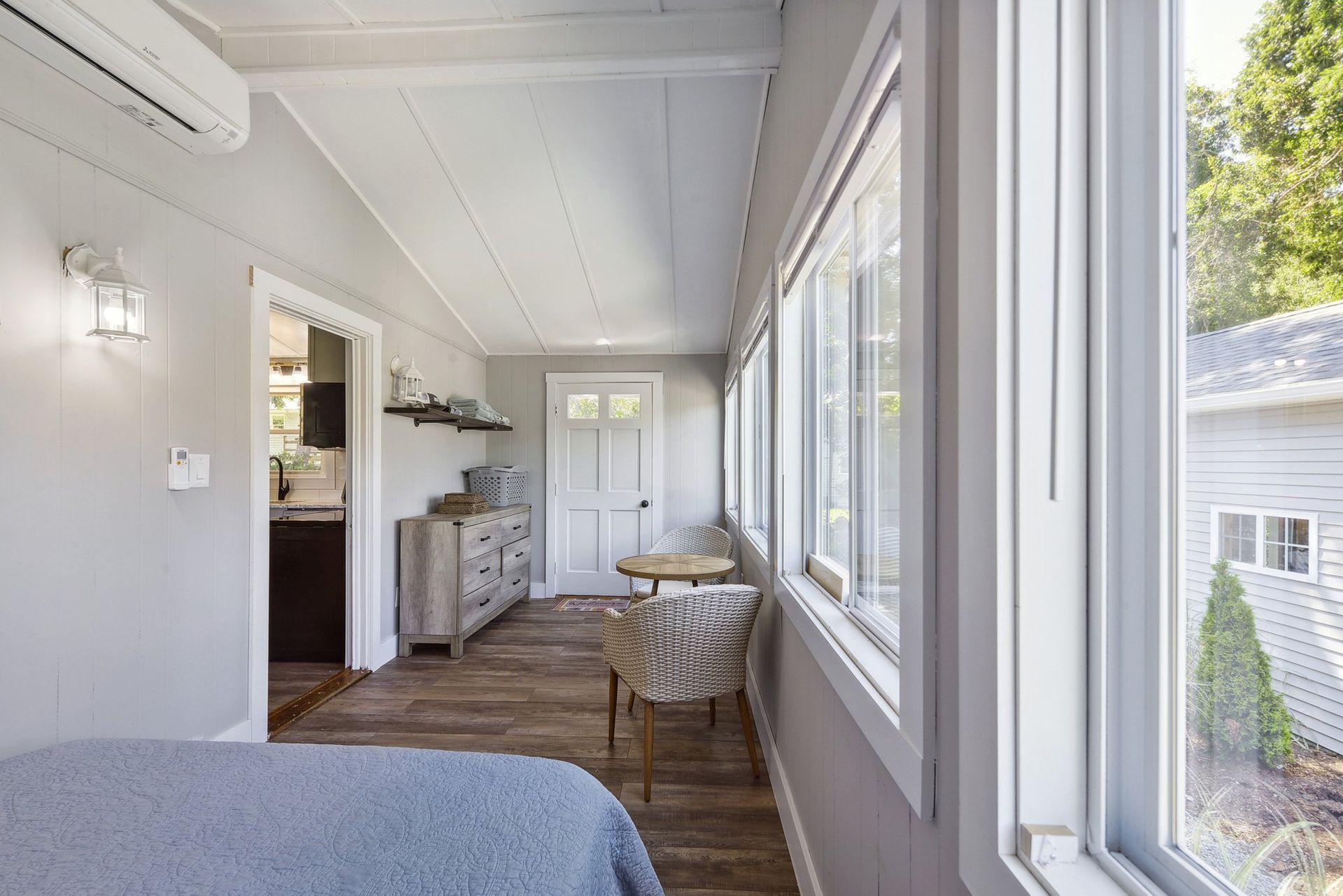 A light, bright sunroom with wood floors, a patterned chair, a small table, a dresser, and a large window view.