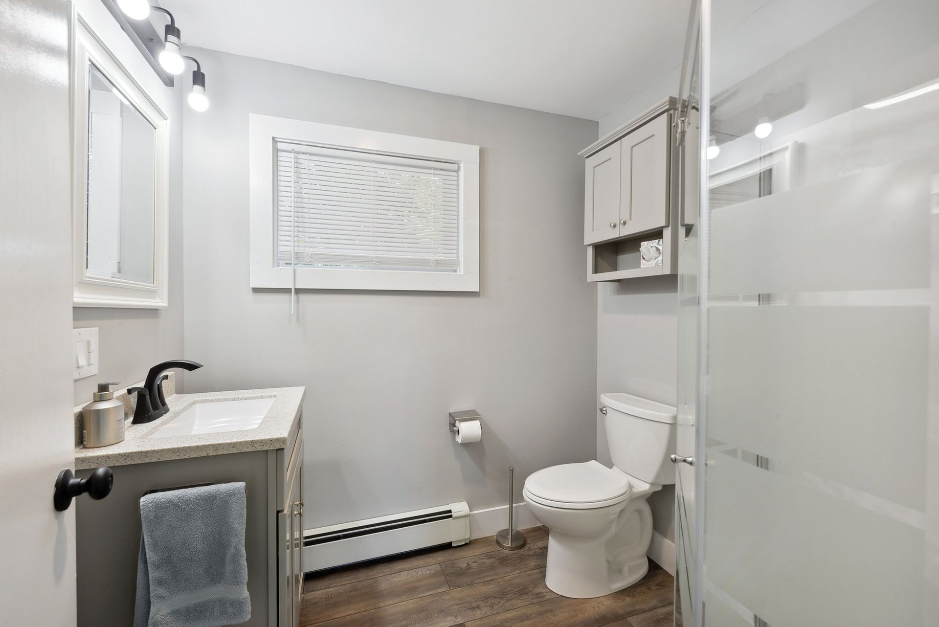 A small, modern gray-toned bathroom featuring a vanity, toilet, and a glass-enclosed shower with wood-look flooring.