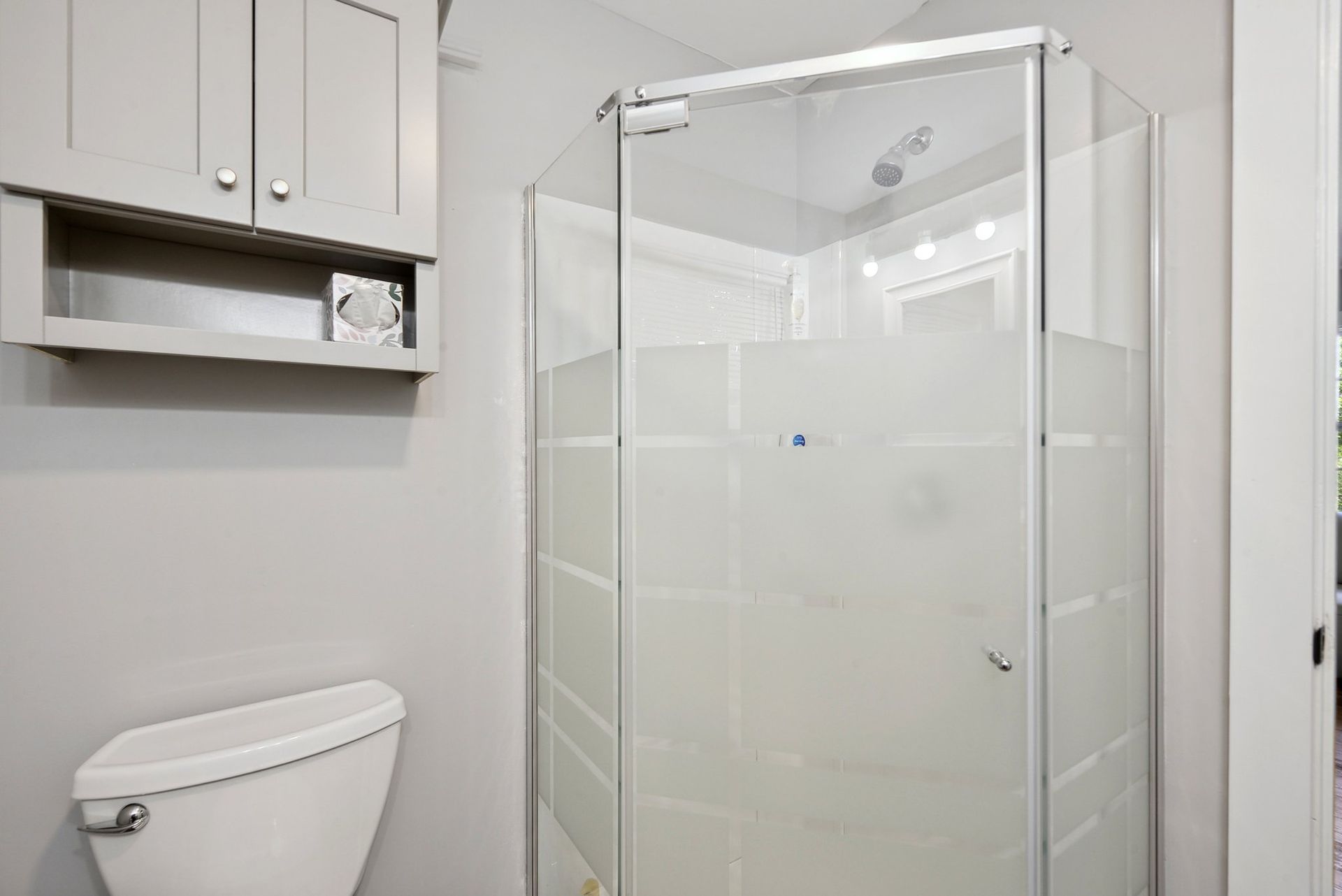 A light gray bathroom featuring a white toilet, a matching wall-mounted storage cabinet, and a corner glass shower stall.