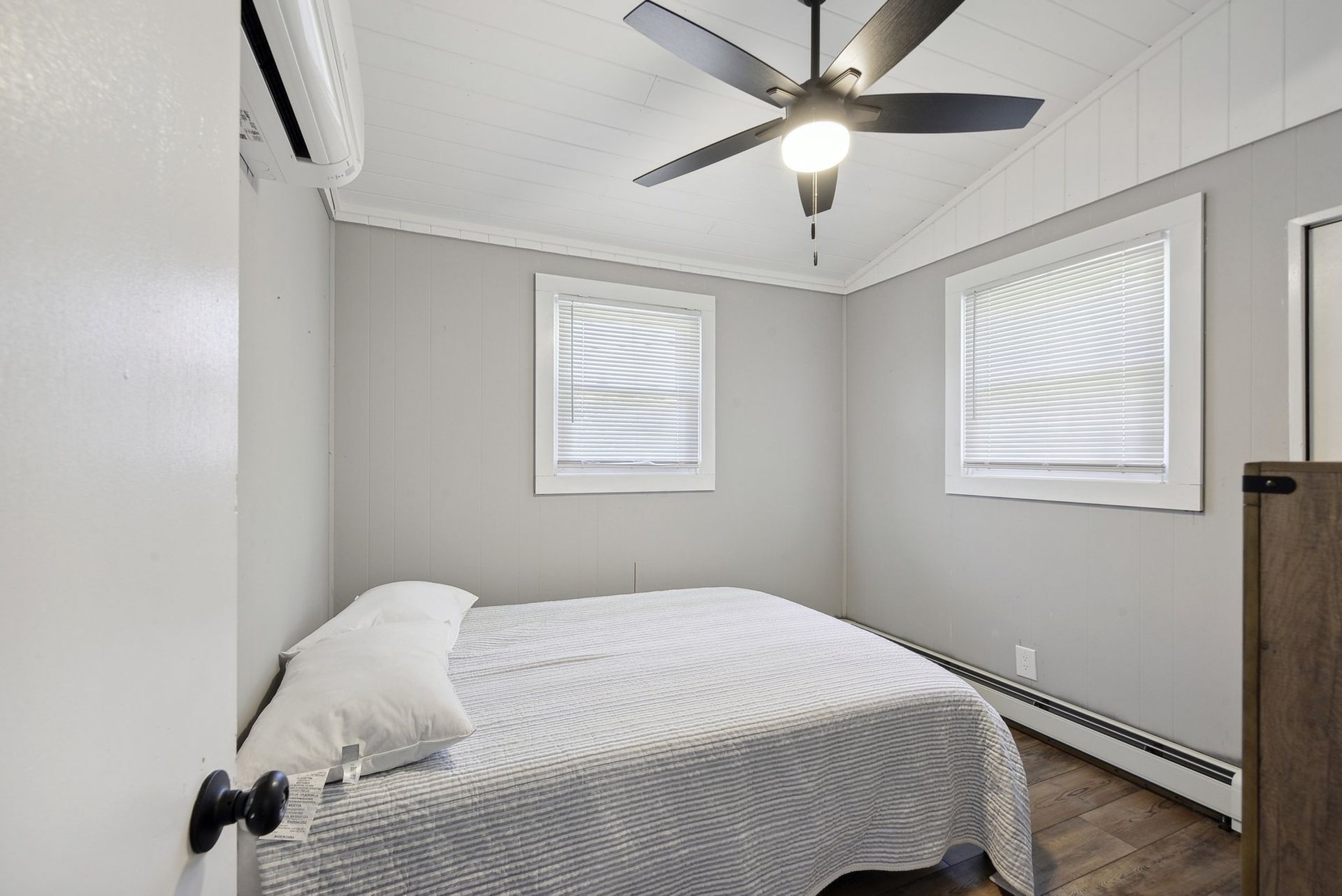 A small, minimalist bedroom with light gray walls, a bed featuring a patterned white spread, and a ceiling fan.