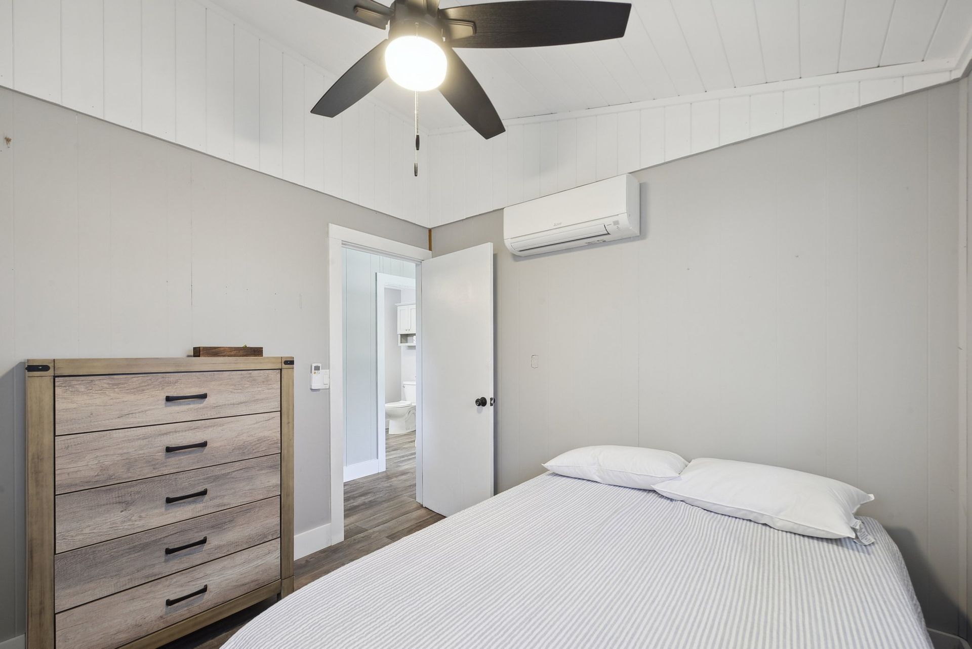 A light-gray bedroom with a white ceiling, a wooden dresser, a ceiling fan, a wall-mounted AC unit, and a made bed.
