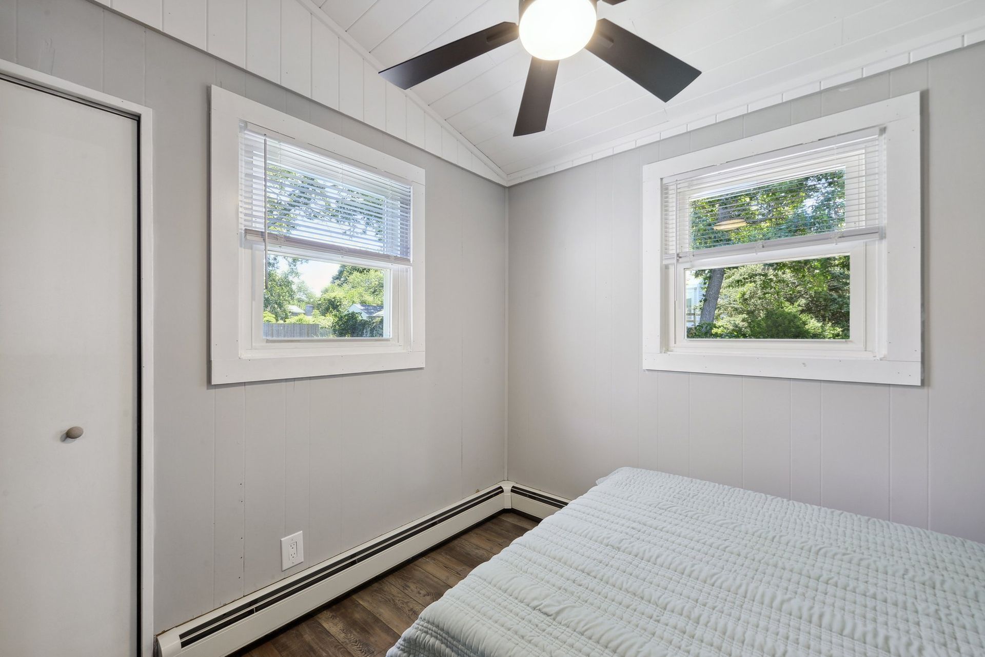 A bedroom with light gray walls, two windows with white blinds, a ceiling fan, and a bed with a light blue textured quilt.