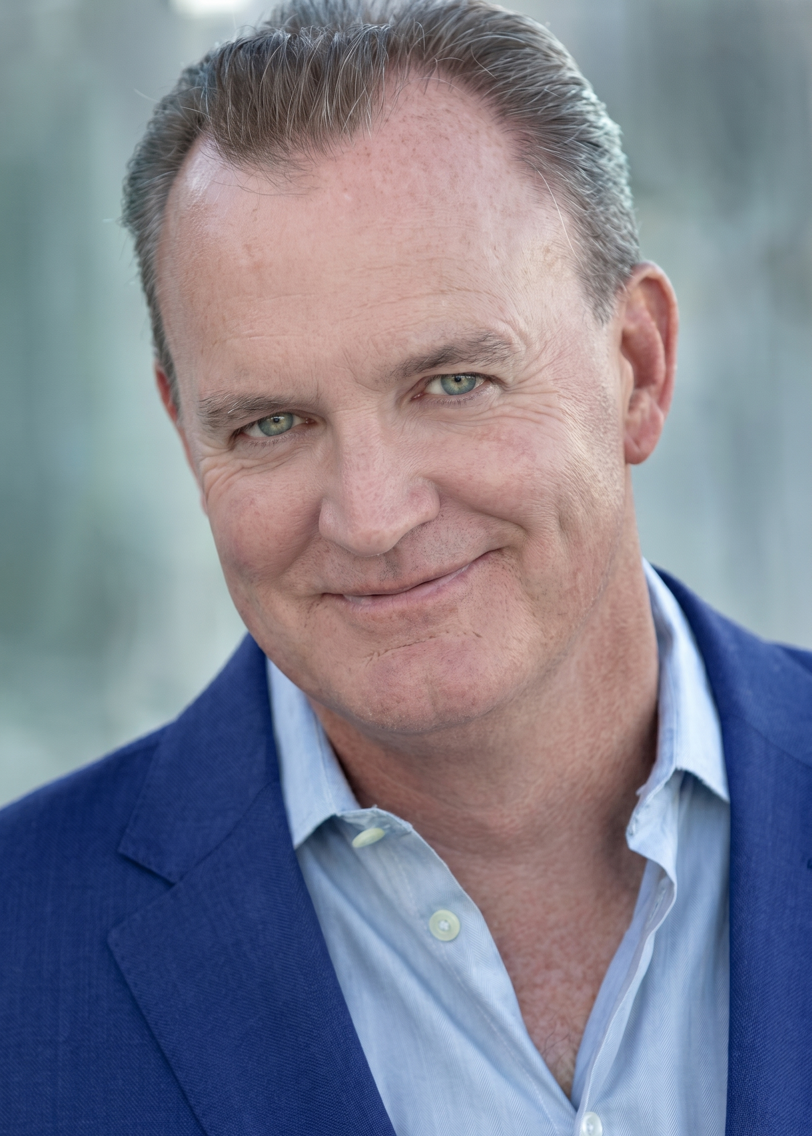 A headshot of a person with short brown hair, wearing a blue blazer and light blue collared shirt, smiling at the camera.
