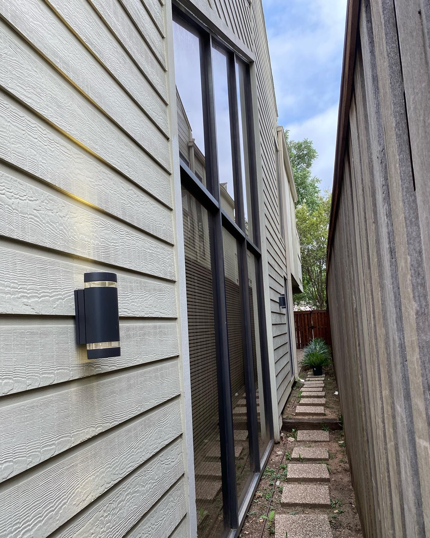 Exterior view of a building with a window and narrow pathway. Light-colored siding and a black light fixture are visible.